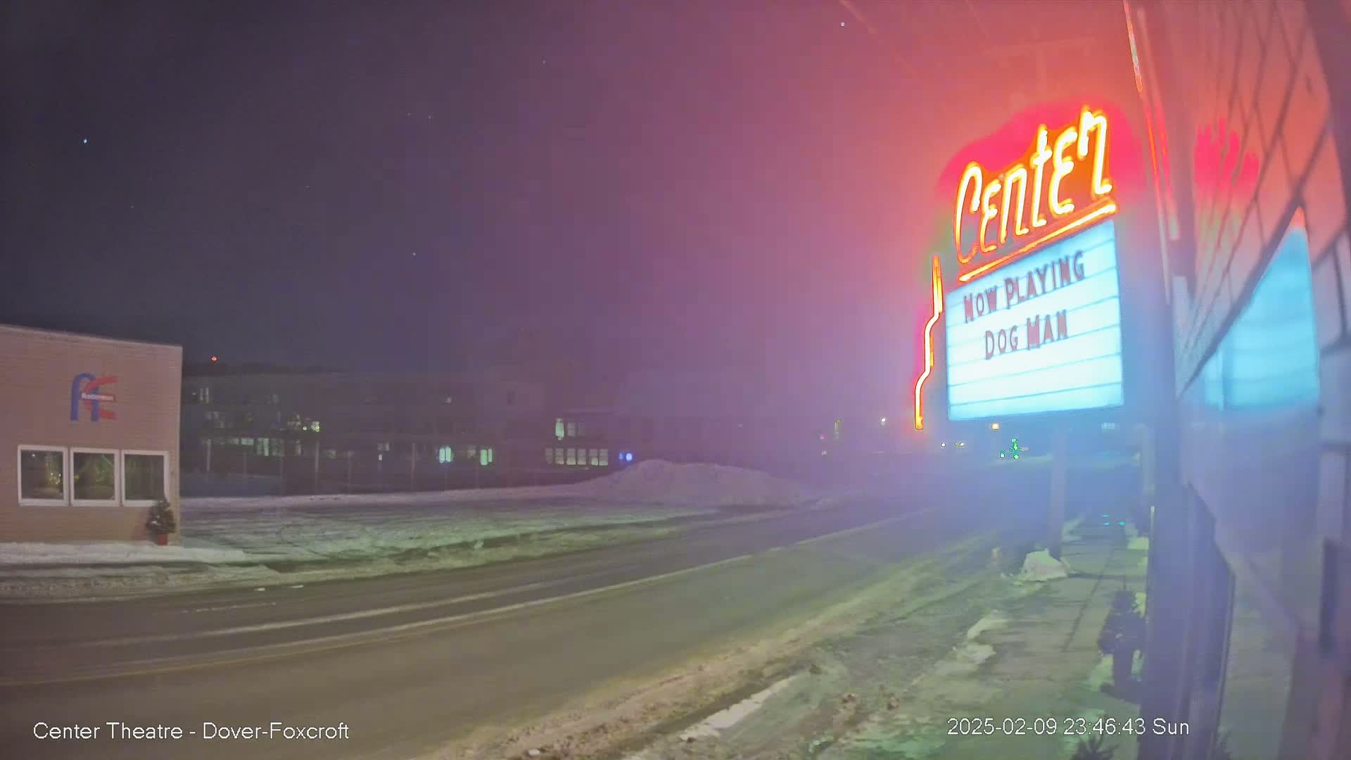 A snow-covered road at night, with a movie theater marquee displaying "Dog Man" under hazy conditions.