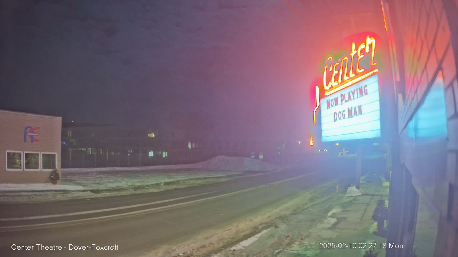 A foggy night scene shows a movie theater marquee advertising "Dog Man," with a snow-covered street and buildings in the background.