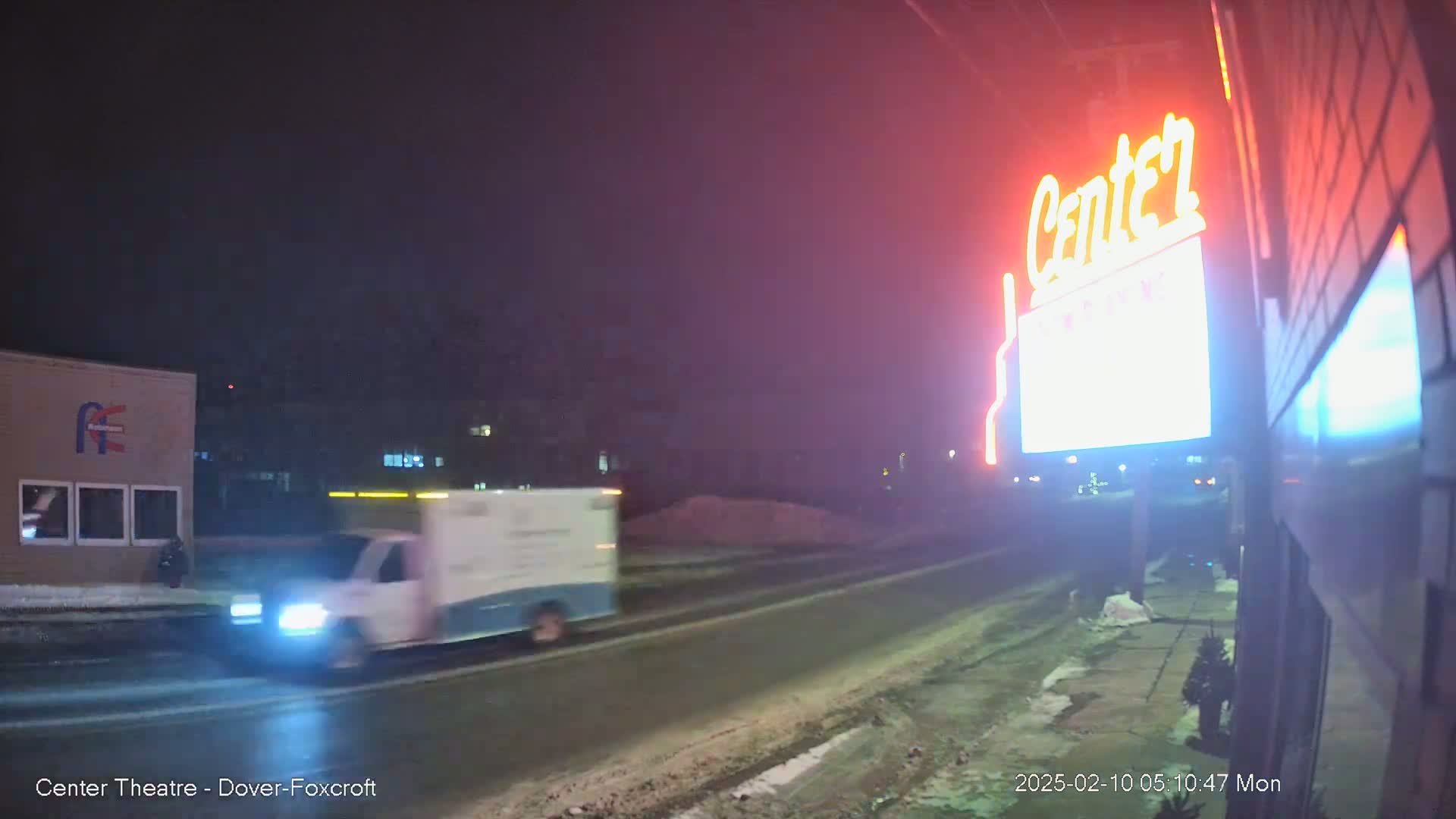 A white van drives past a brightly lit theater sign on a snowy night.