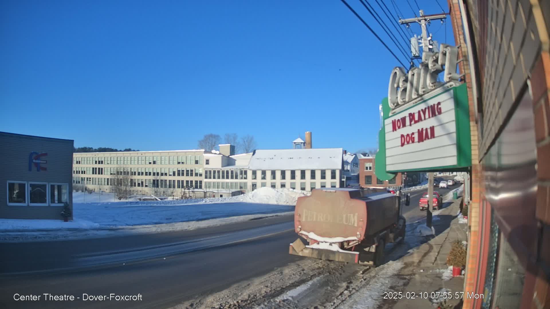 A petroleum tanker truck is parked on a snow-covered street in front of a movie theater and several buildings under a clear, sunny sky.