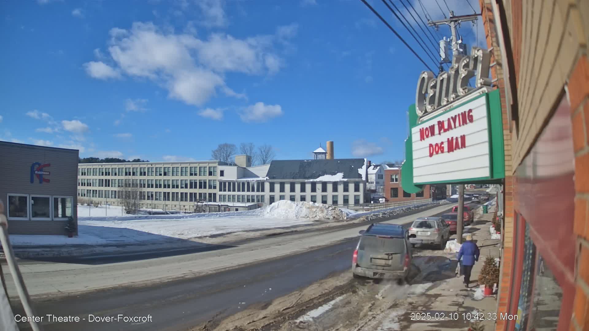 A sunny winter day shows a street scene with cars parked along the side, snow on the ground, and several buildings in the background.