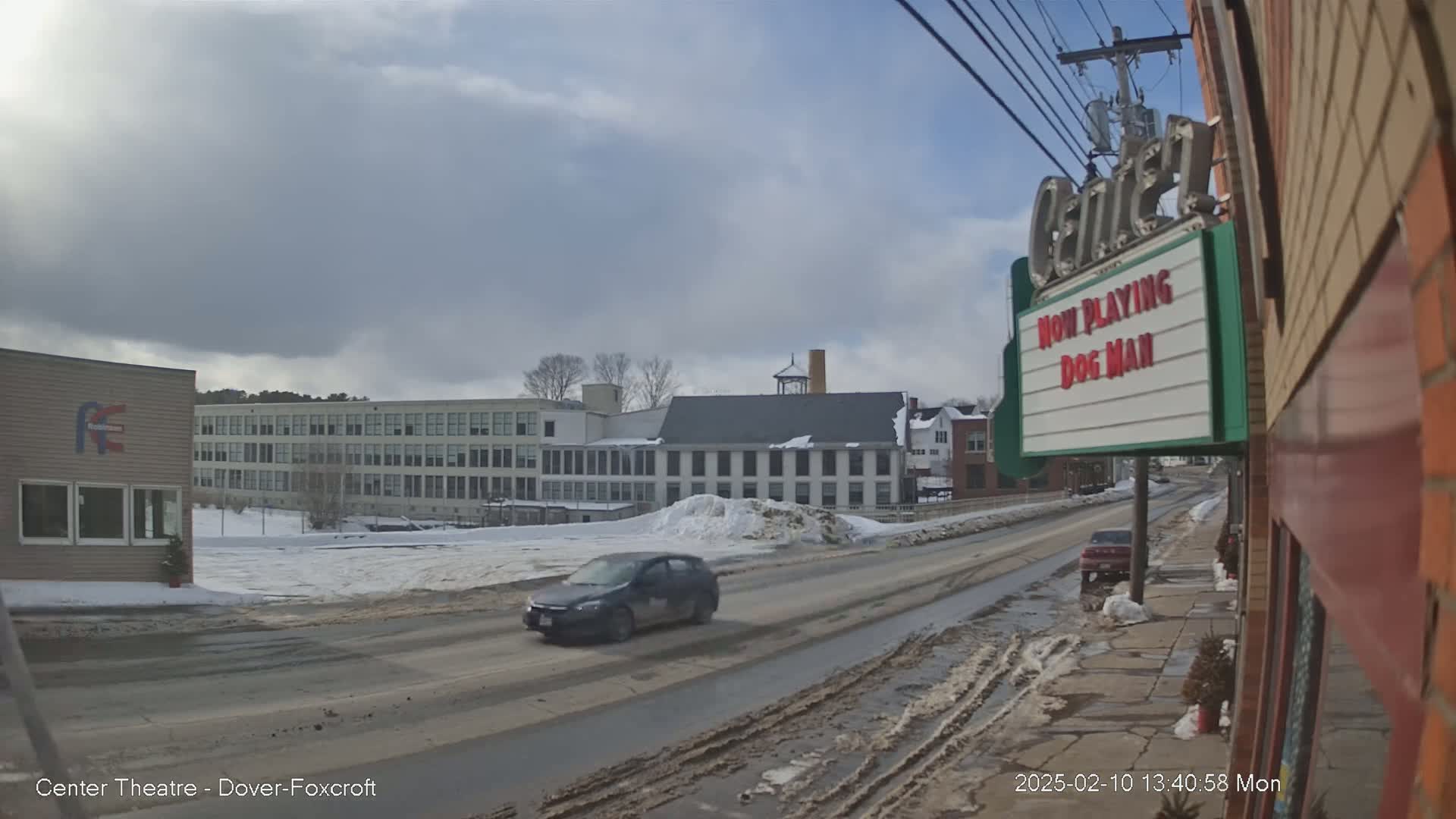 A mostly cloudy day shows a snow-covered street scene with a car driving past buildings, including a large factory and a movie theater marquee.