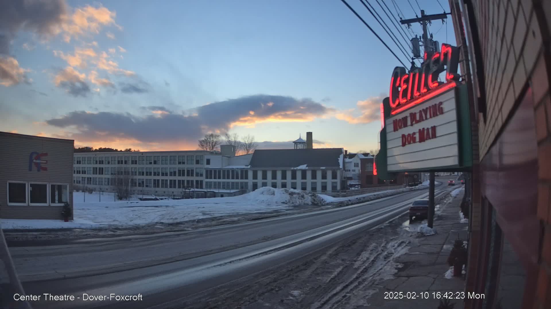 A snow-covered street scene at sunset shows a multi-story building and a movie theater marquee displaying "Dog Man."