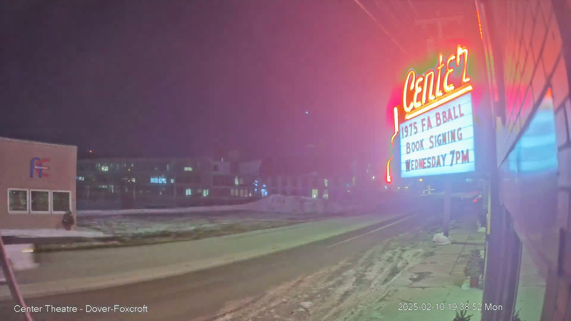 A nighttime street scene shows a snow-covered road in front of a building with a neon sign advertising a book signing, under a hazy, dark sky.