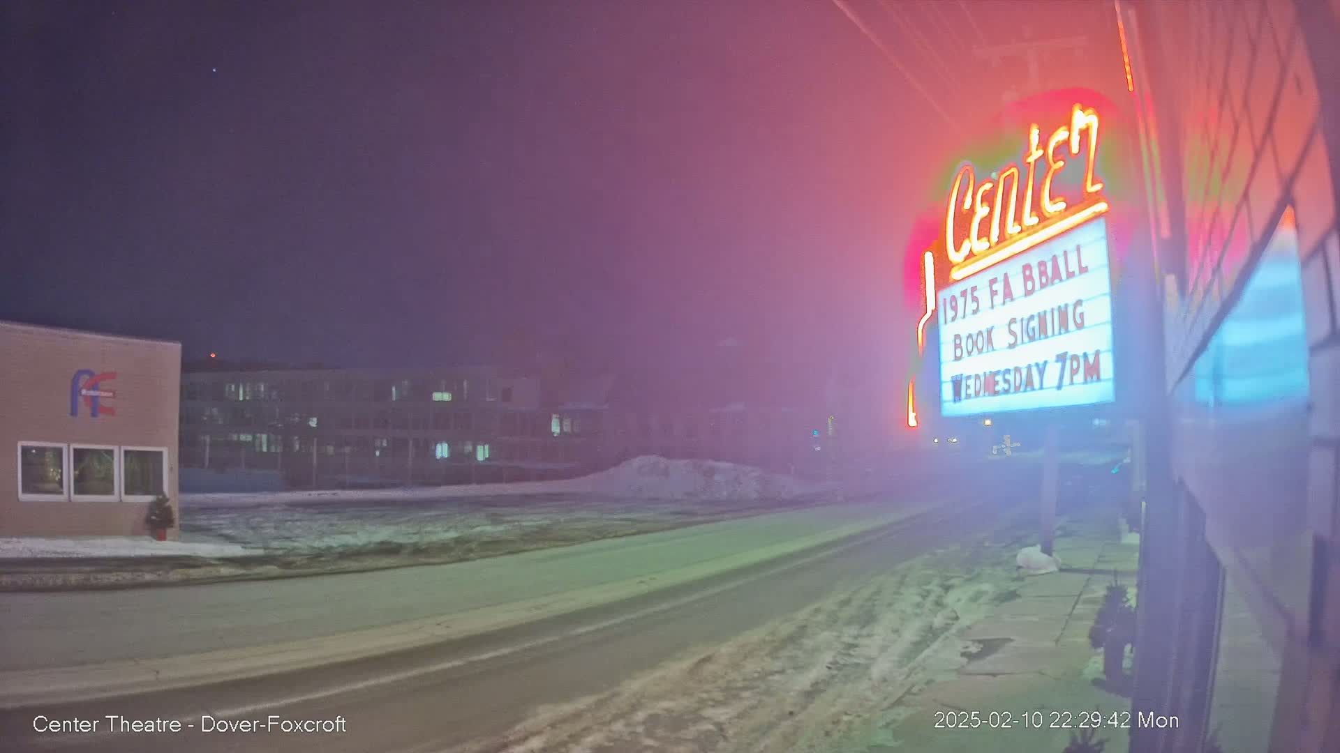 A snow-covered street at night is seen in front of a theater with a marquee advertising a book signing, under a hazy sky.