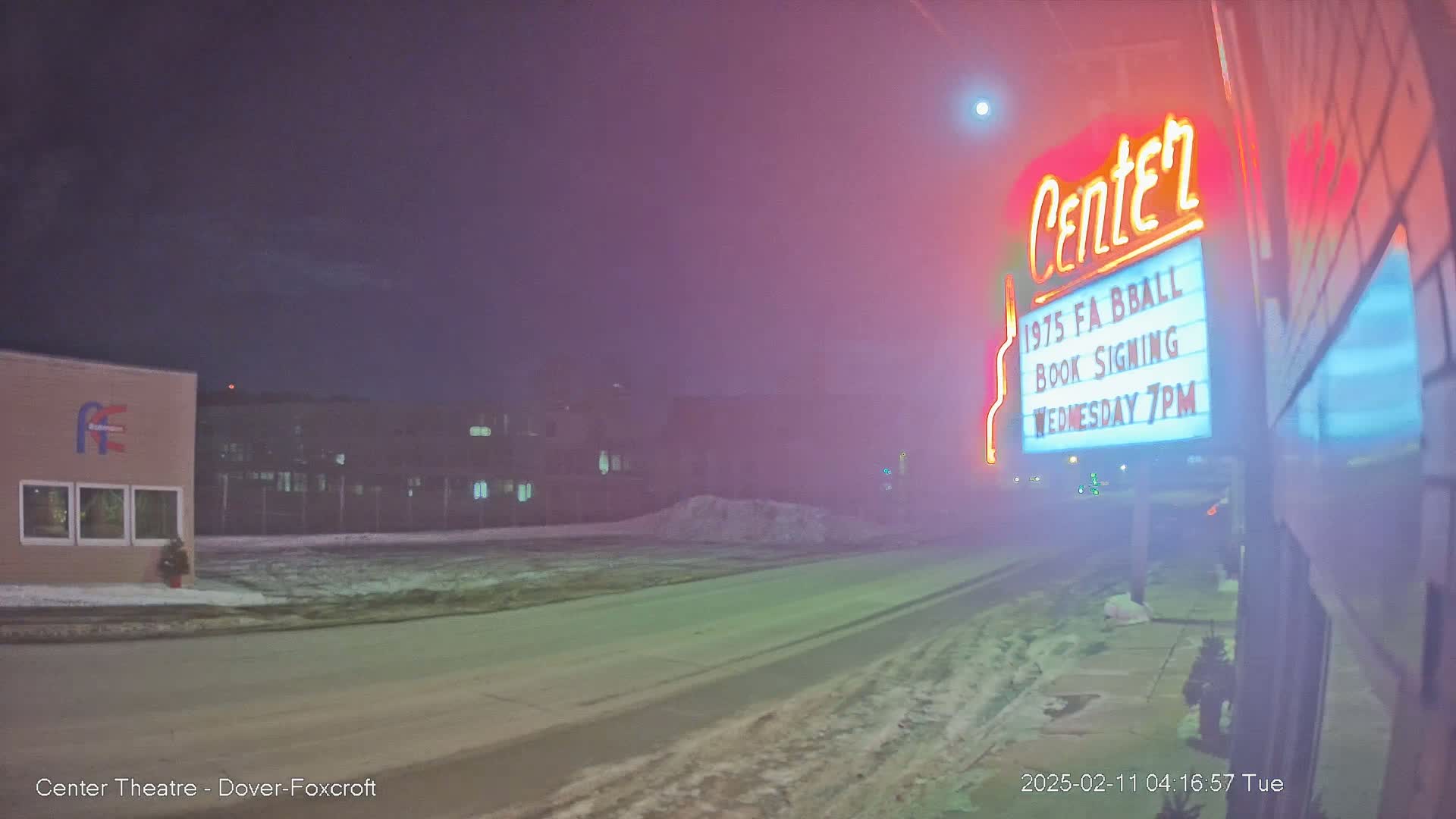 A foggy night scene shows a street with snow on the ground, a building with a neon sign advertising a book signing, and another building in the distance.