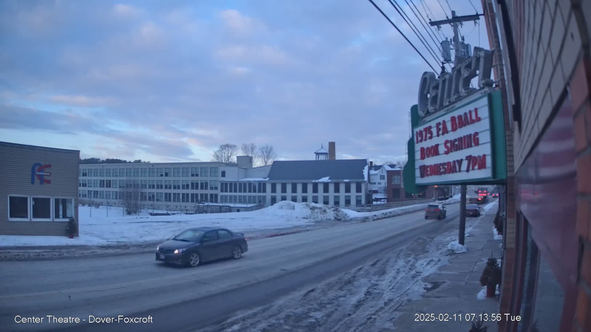 A snow-covered street scene shows several buildings, including a large multi-story building and a smaller one with a marquee, under a partly cloudy sky.