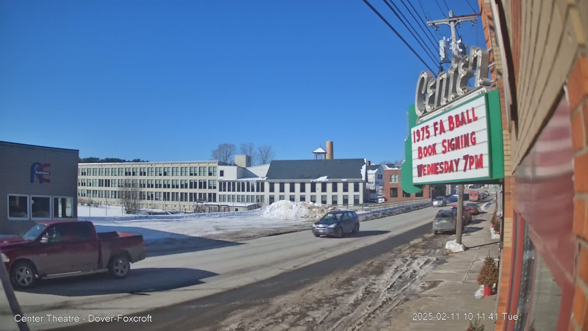 A sunny winter day shows a street scene with several vehicles, some snow on the ground, and multi-story buildings in the background.