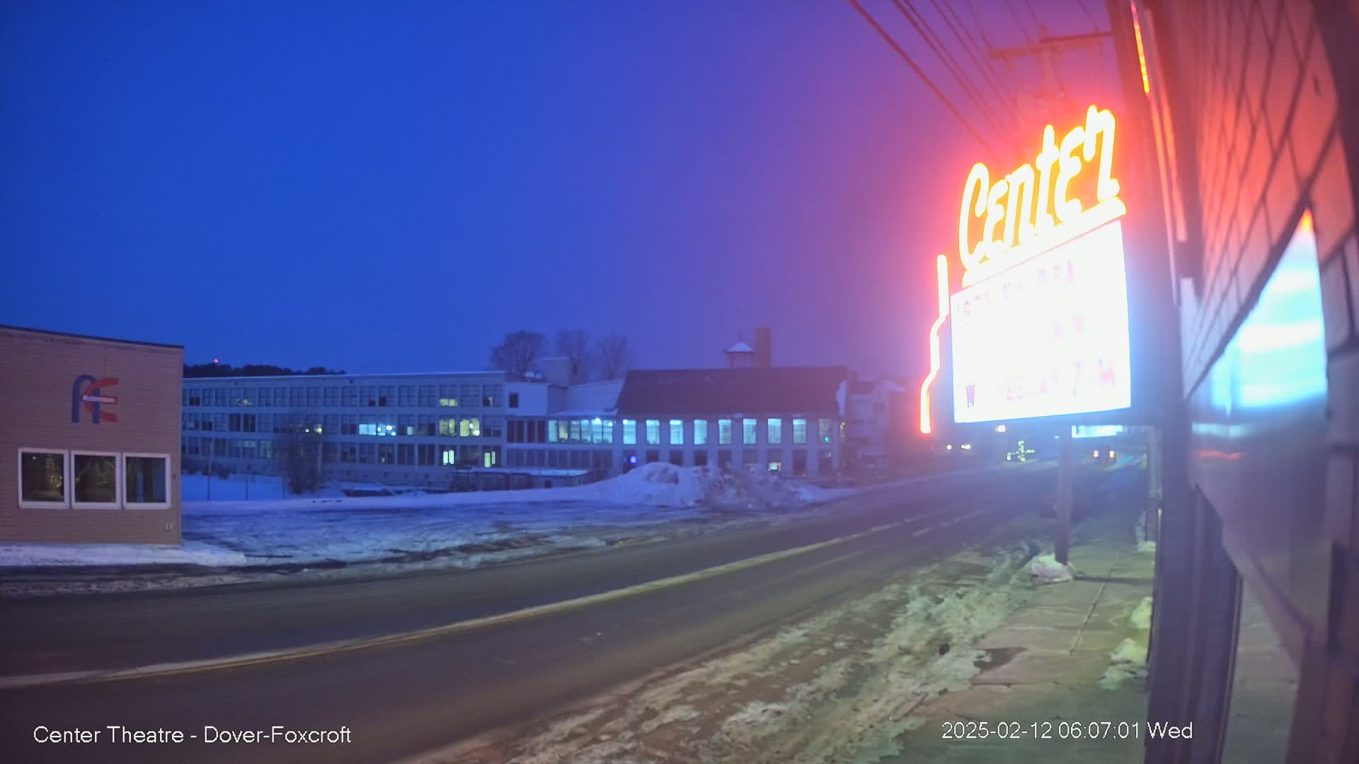 A snow-covered street scene at twilight shows a neon sign for a theater, a multi-story building, and a smaller building in the background.