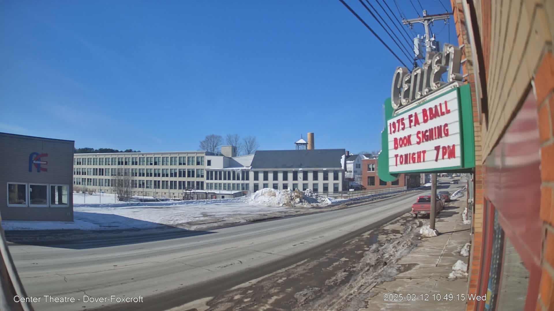 A street scene on a sunny winter day shows a multi-story building, smaller buildings and a snow-covered empty lot, with cars parked along the road.