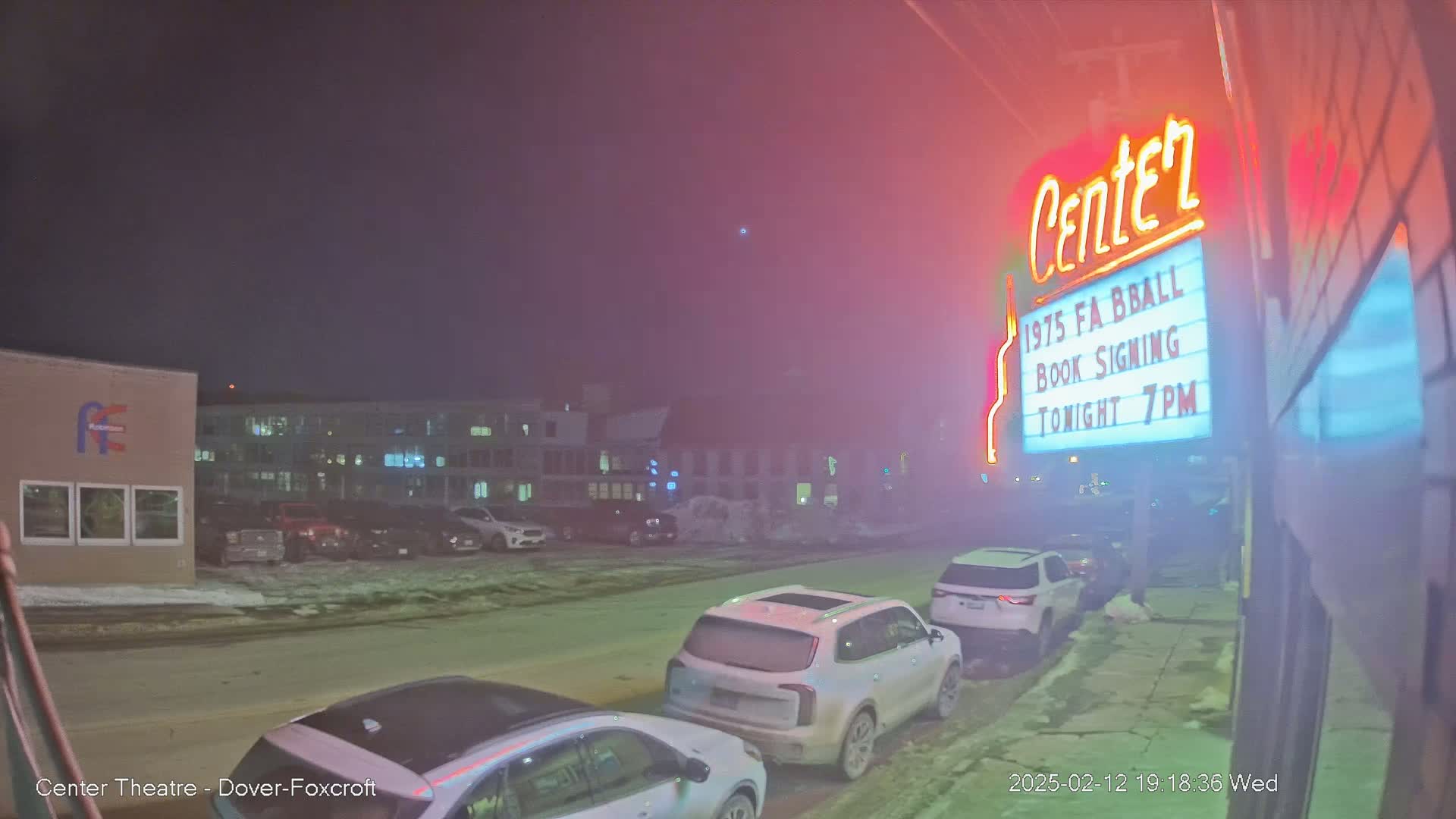 A nighttime scene of a snow-covered street with parked cars in front of a building displaying a marquee advertising a book signing.