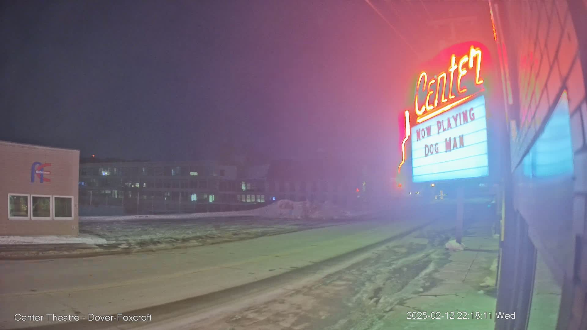 A foggy night scene shows a movie theater marquee advertising "Dog Man," with a snow-covered street and buildings in the background.