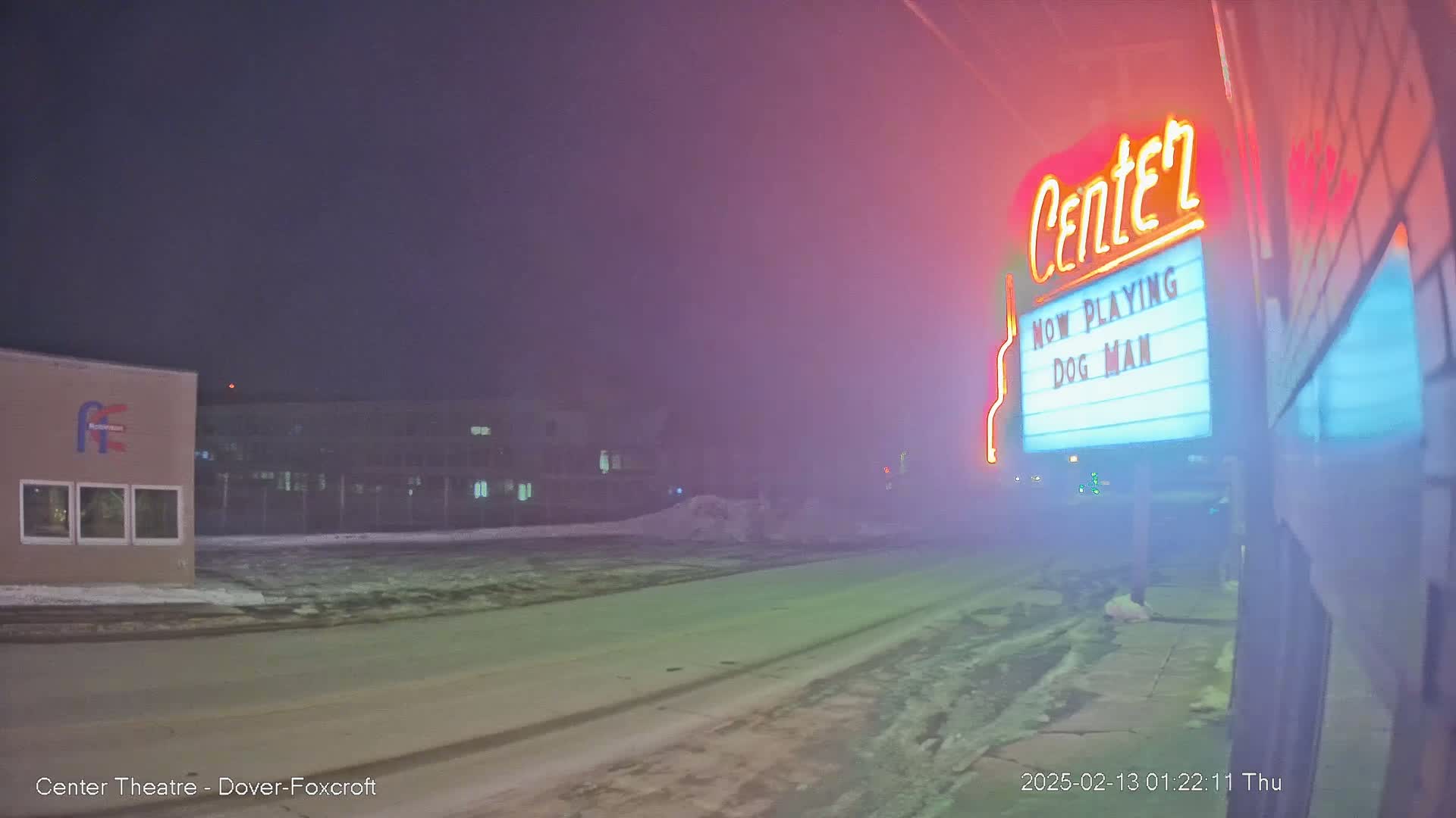 A foggy night scene shows a movie theater marquee advertising "Dog Man," with a snow-covered street and buildings in the background.