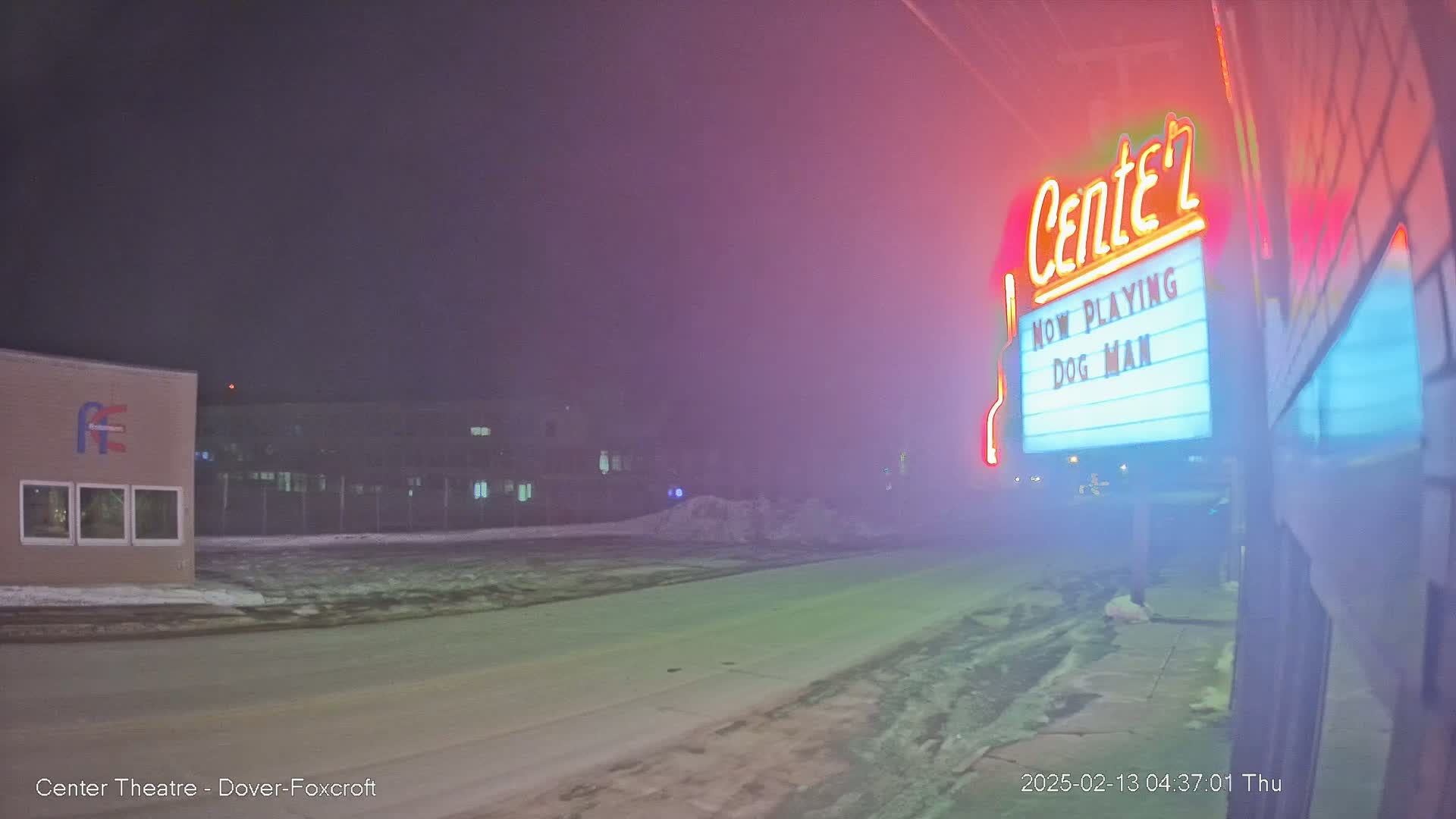 A foggy night scene shows a movie theater marquee advertising "Dog Man," with a snowy road and buildings in the background.