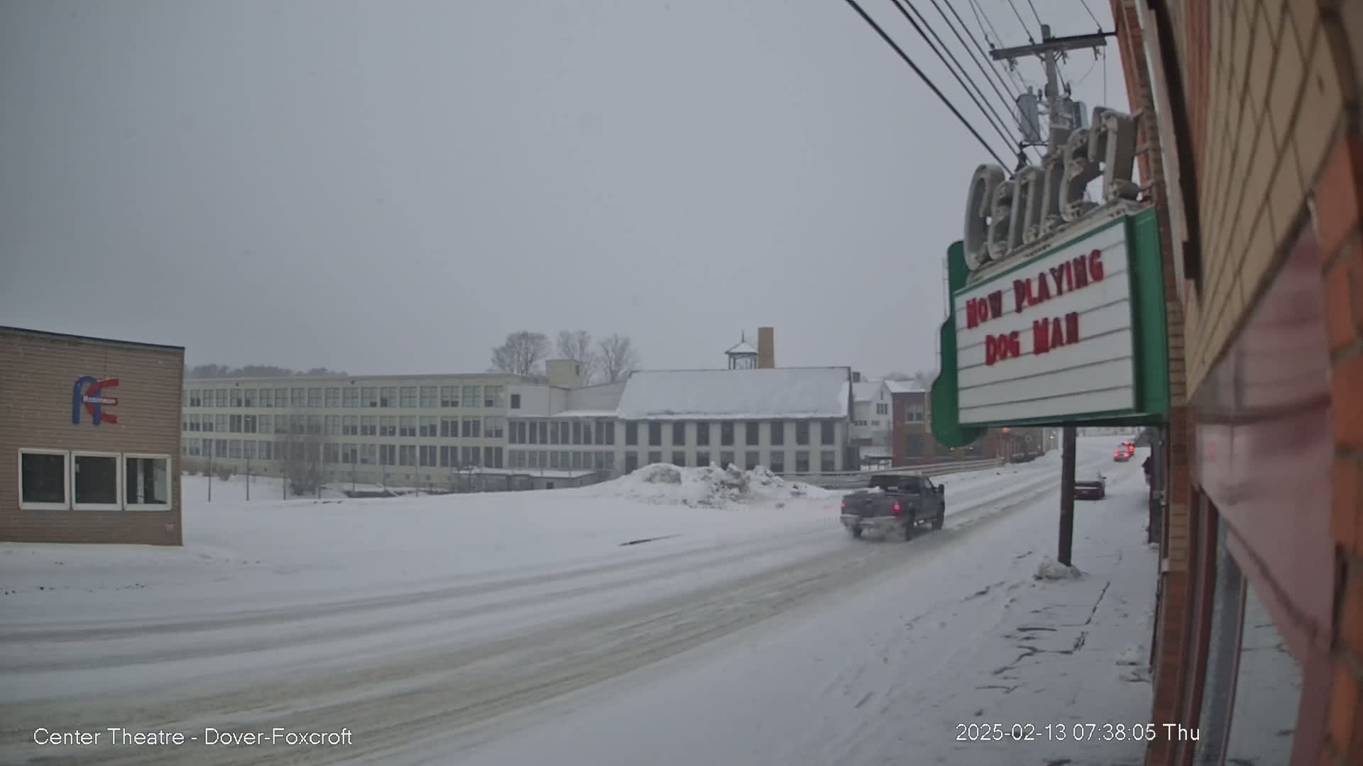 A snow-covered street scene shows a pickup truck driving past buildings, including a large school and a movie theater marquee advertising "Dog Man," under a heavy, overcast sky.
