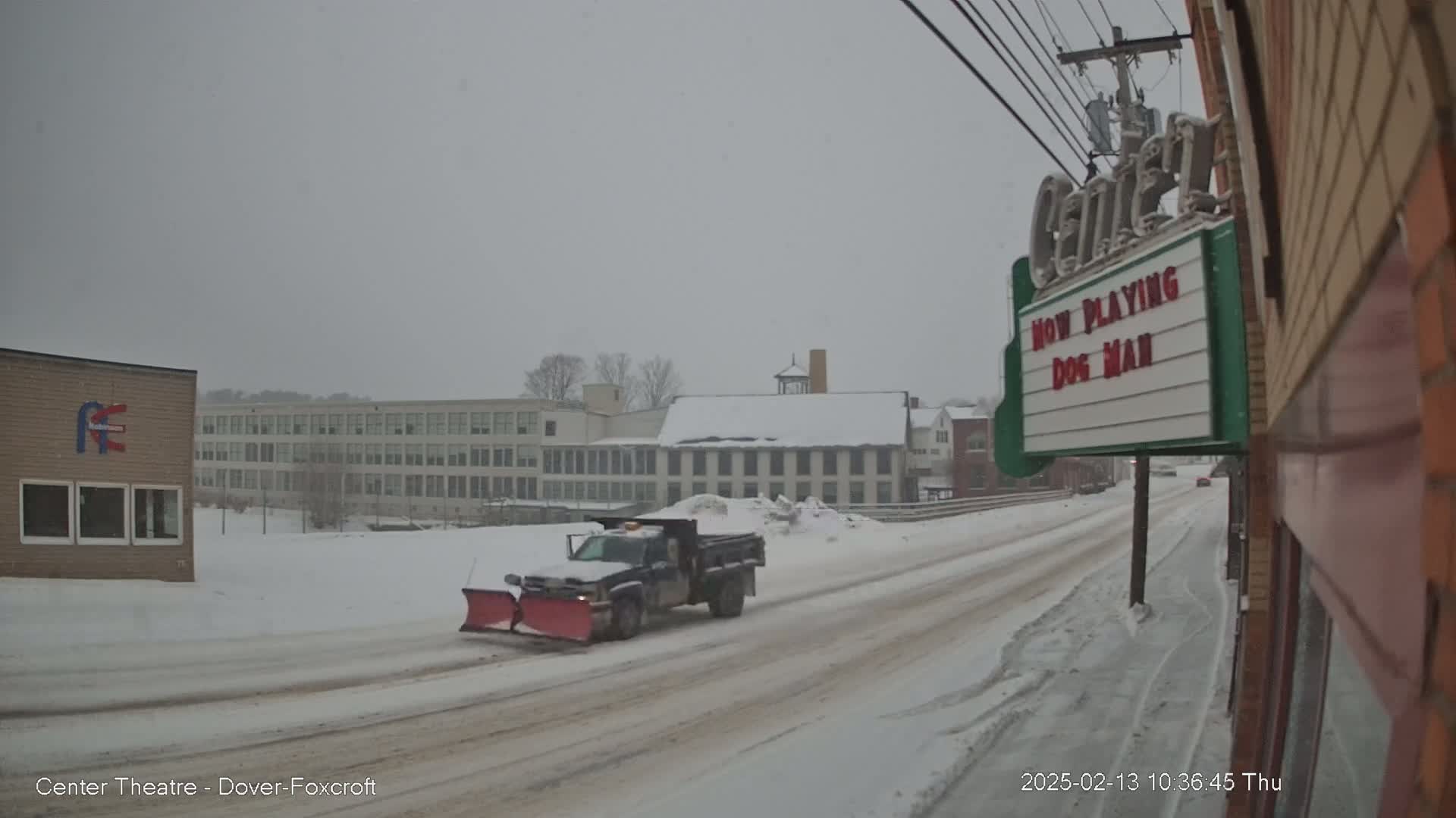 A snow-covered street scene shows a snowplow clearing a road in front of buildings, with light snow falling.