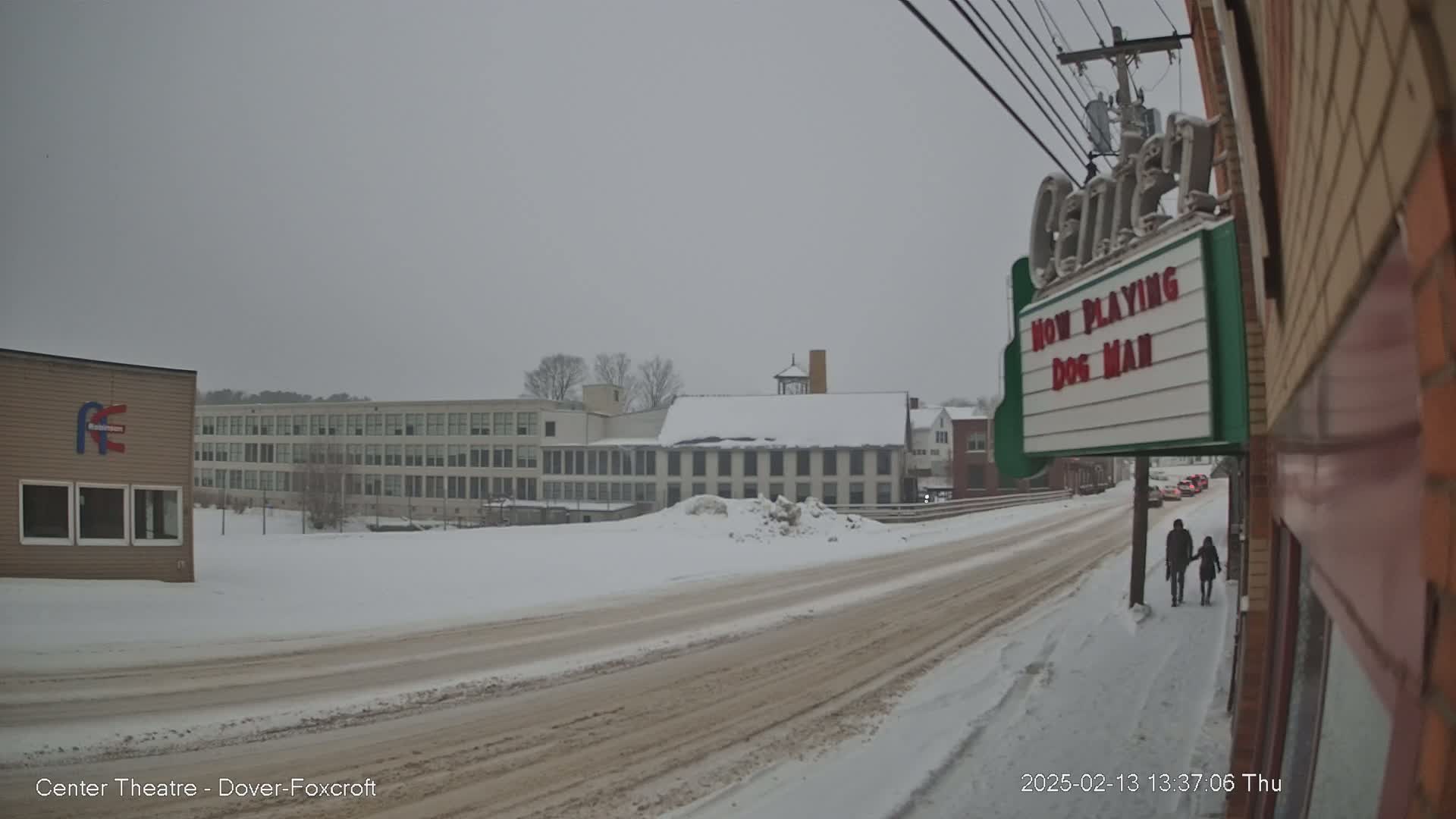 A snow-covered street scene shows a couple walking hand-in-hand past a movie theater marquee under an overcast sky.