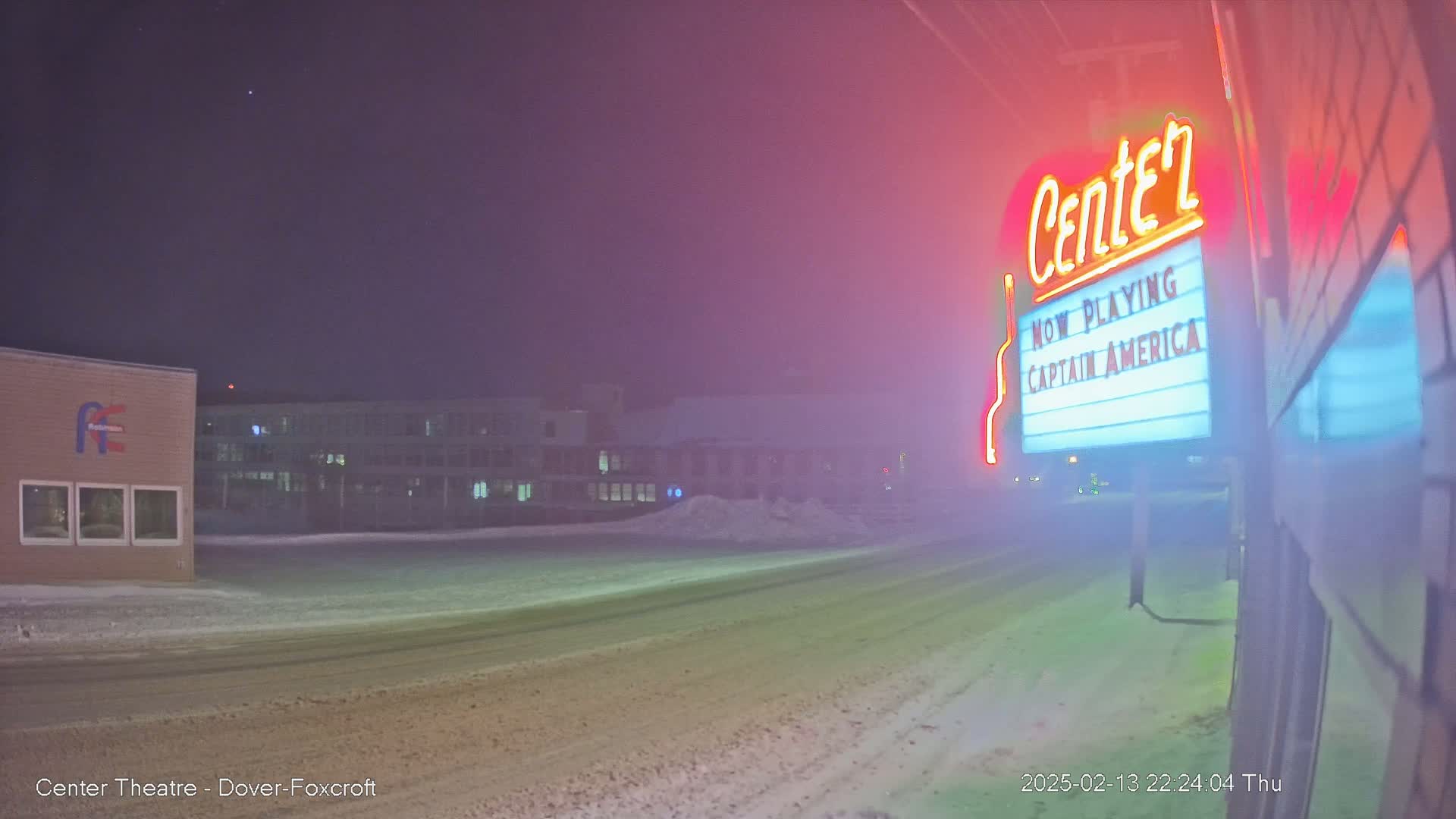 A snow-covered street at night is seen, with a movie theater marquee advertising "Captain America" illuminated in the fog.