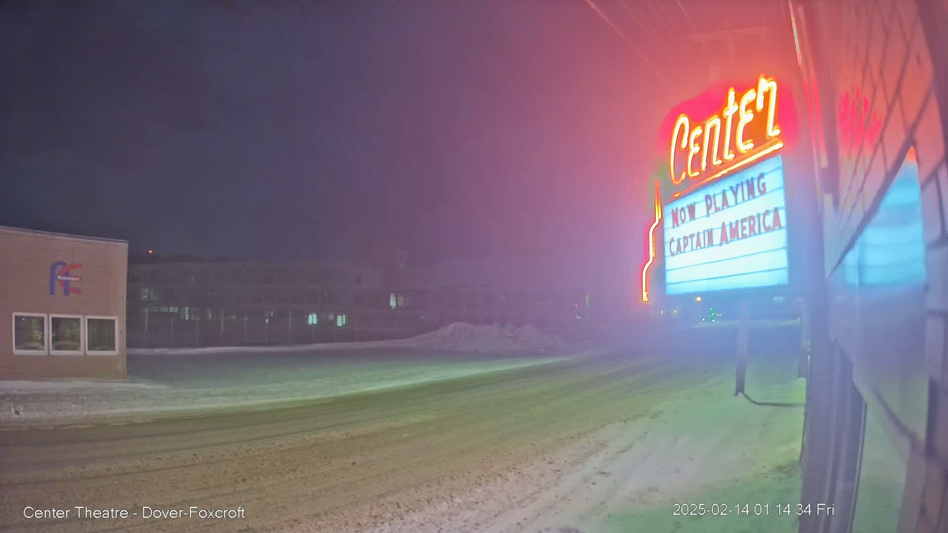 A snow-covered street at night is dimly lit, with a movie theater marquee showing "Captain America" in the foreground and buildings in the background under a foggy sky.