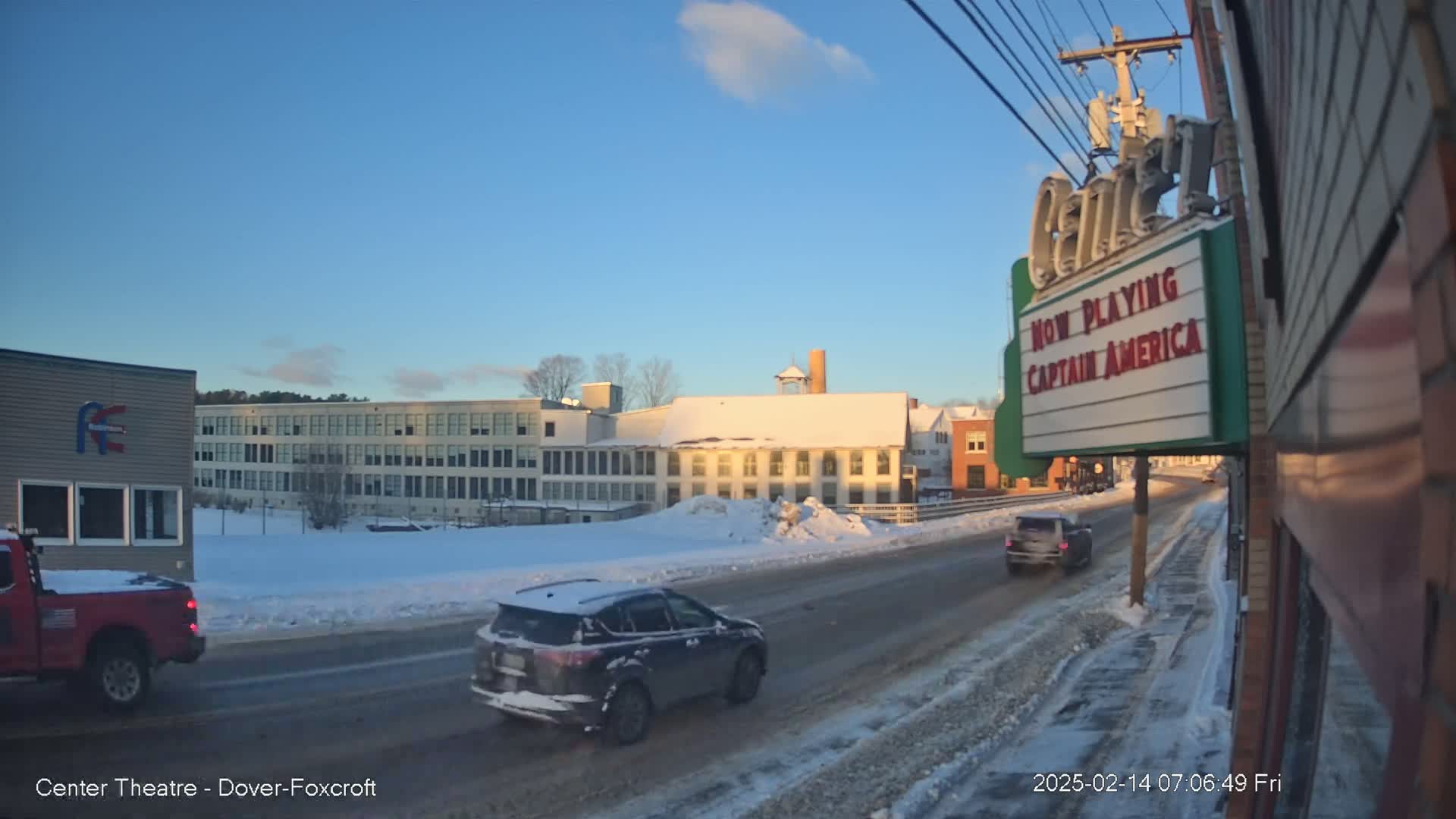 A sunny, snowy street scene shows a few vehicles traveling along a road past a movie theater marquee and several buildings.