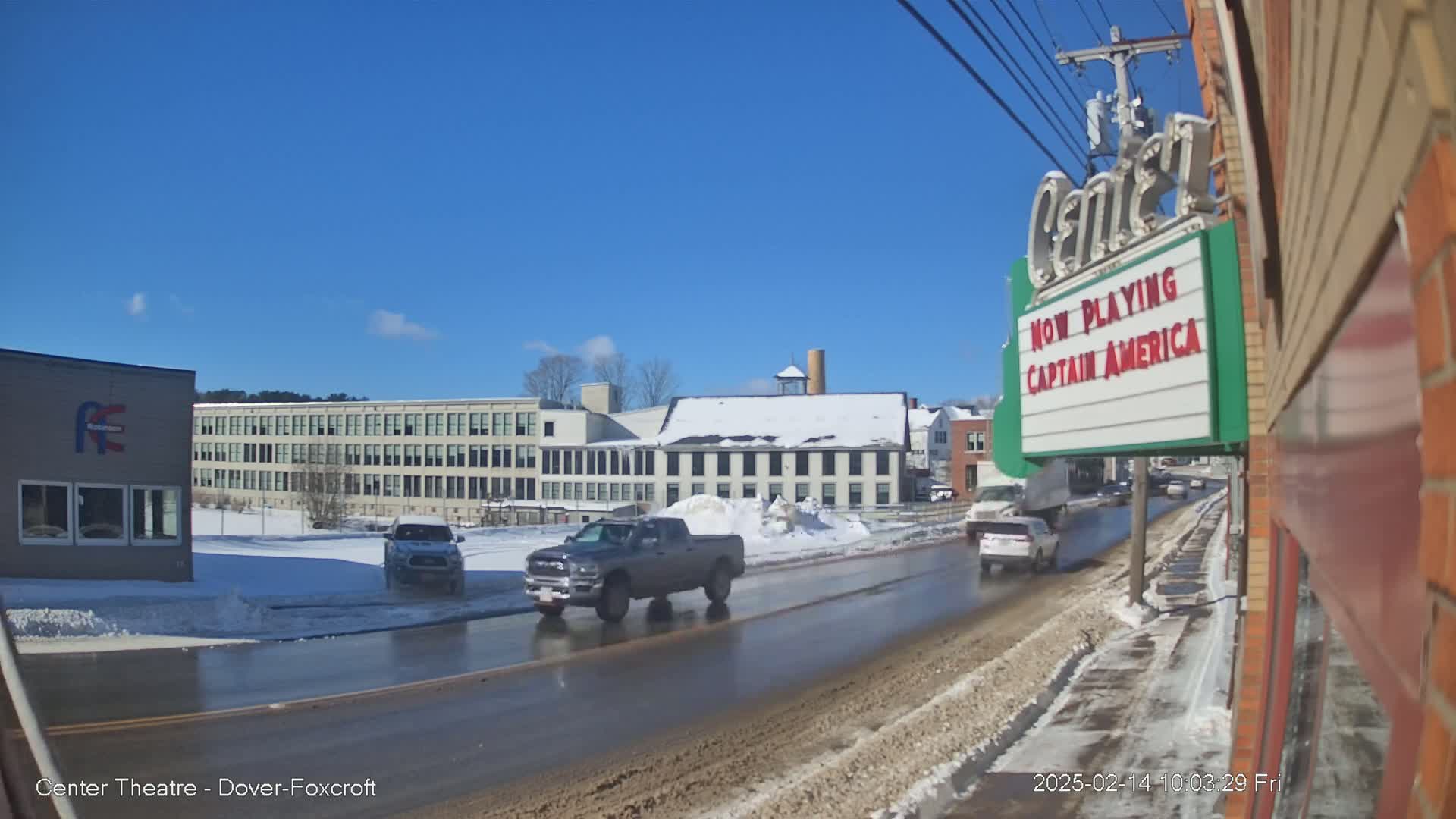 A partly sunny winter day reveals a street scene with several vehicles, including a gray pickup truck, driving past a snow-covered building and a movie theater marquee advertising "Captain America."