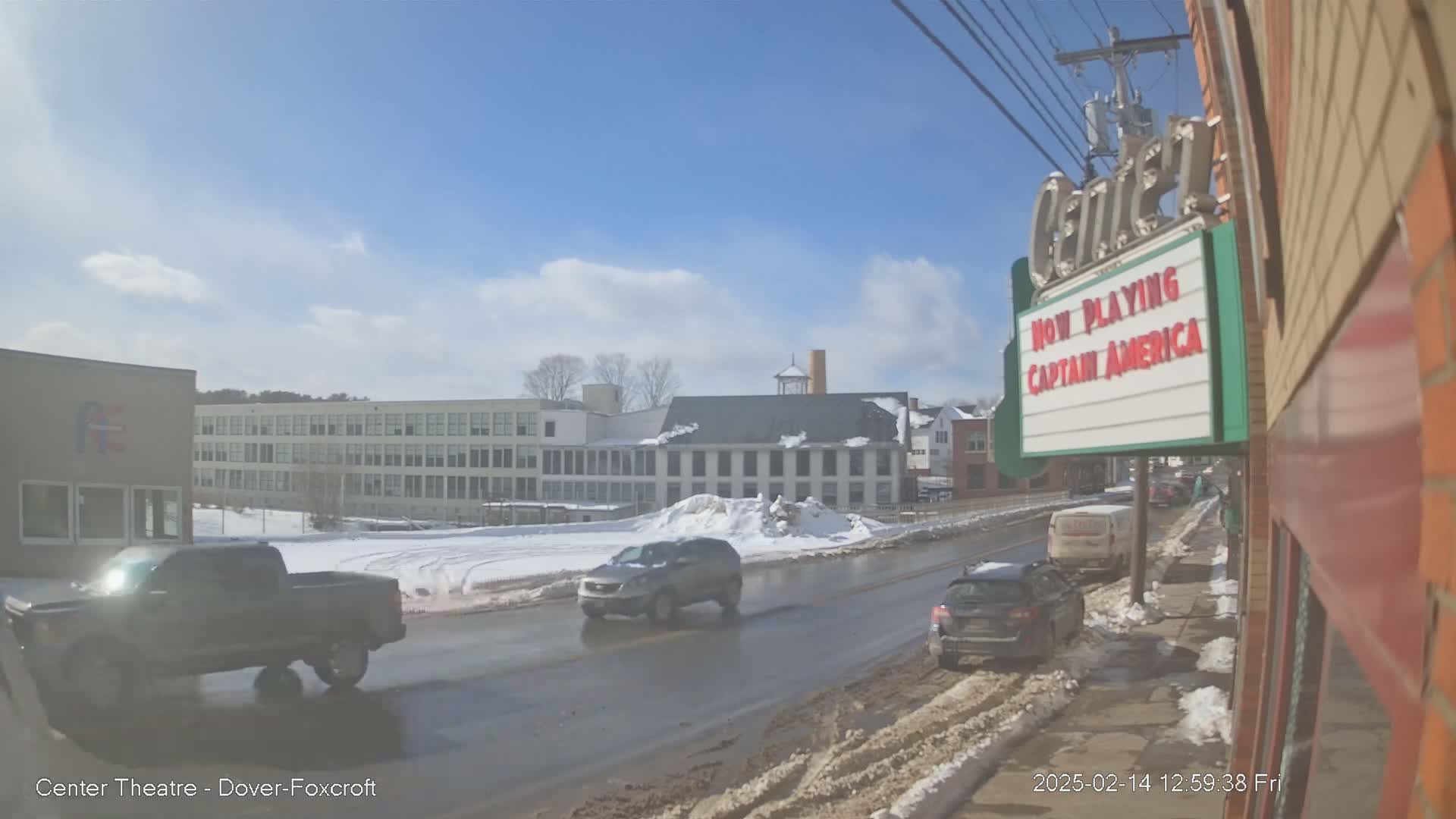 A sunny winter day shows a street scene with several cars driving past a movie theater marquee and multi-story buildings partially covered in snow.
