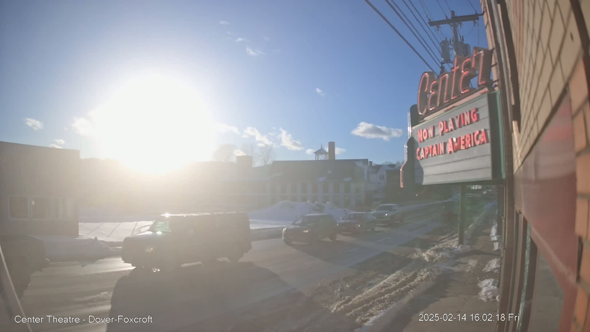 A sunny day with some snow on the ground shows a street scene with several cars driving by a movie theater marquee advertising "Captain America."
