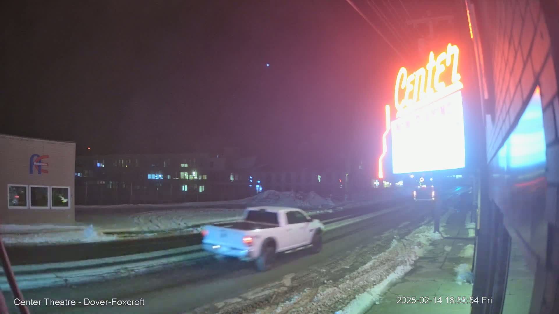 A white pickup truck drives on a snow-covered road at night past a building with illuminated windows and a neon sign.