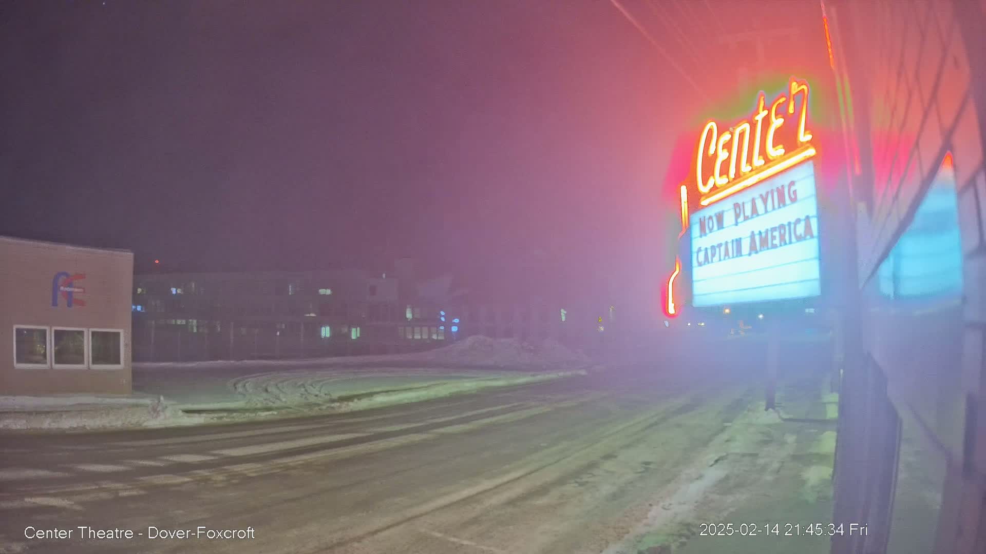A snowy street at night is shrouded in fog, with a movie theater marquee advertising "Captain America" brightly lit in the foreground.