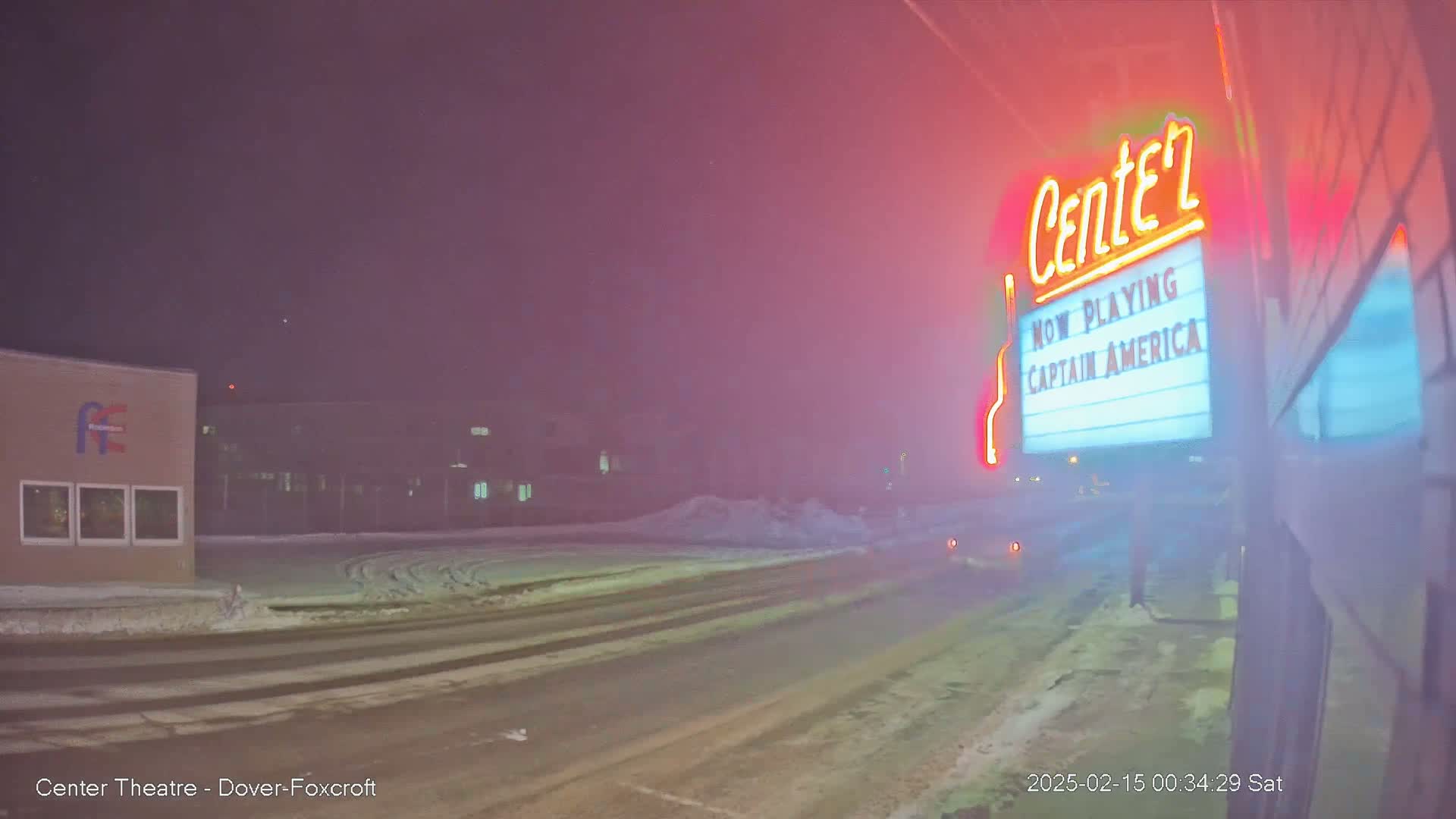 A snow-covered street at night shows a movie theater marquee advertising "Captain America," with a hazy, pinkish glow in the air.