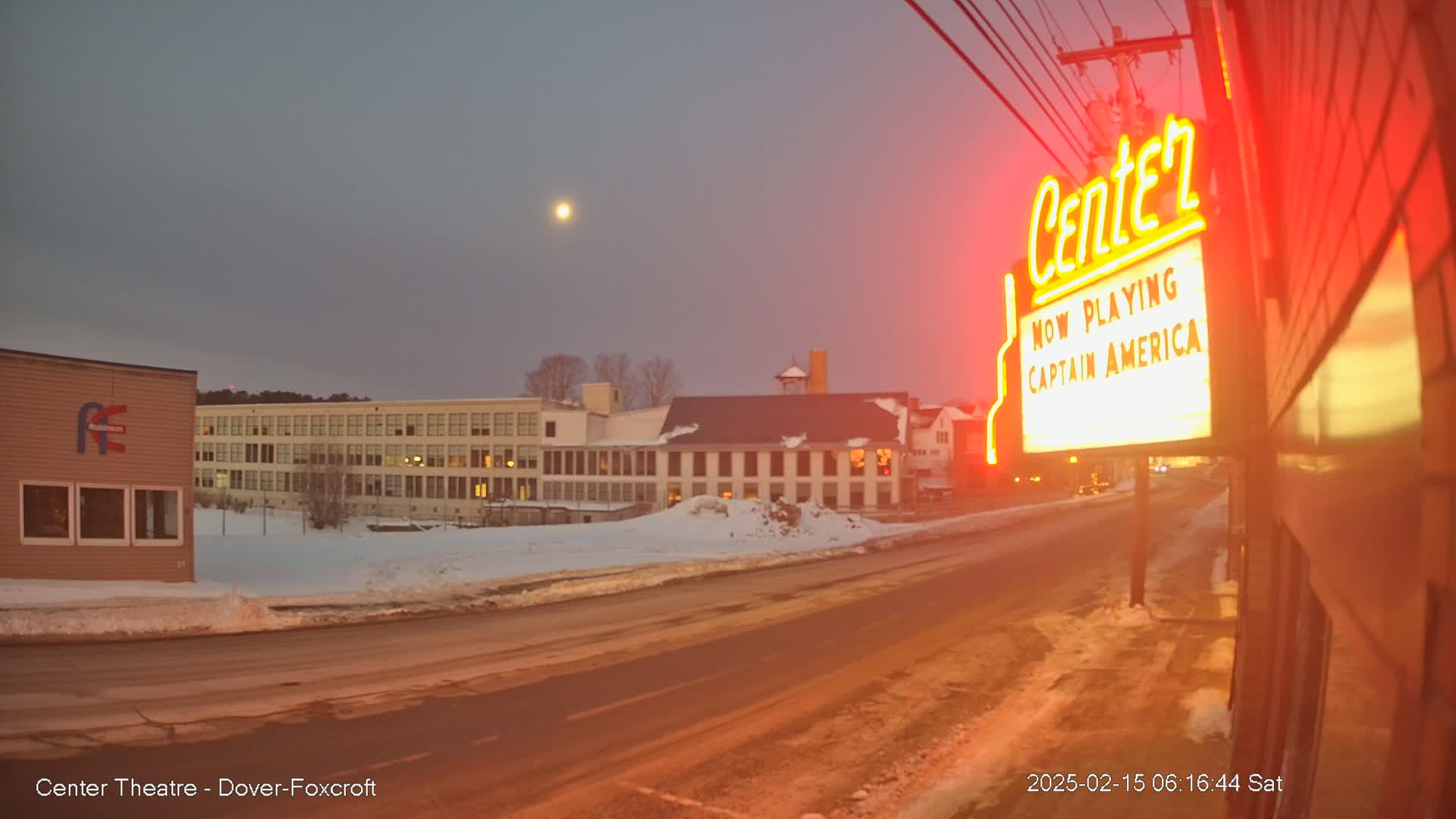 A snow-covered street scene at twilight shows a movie theater marquee advertising Captain America, with a multi-story building and a moon visible in the dusky sky.