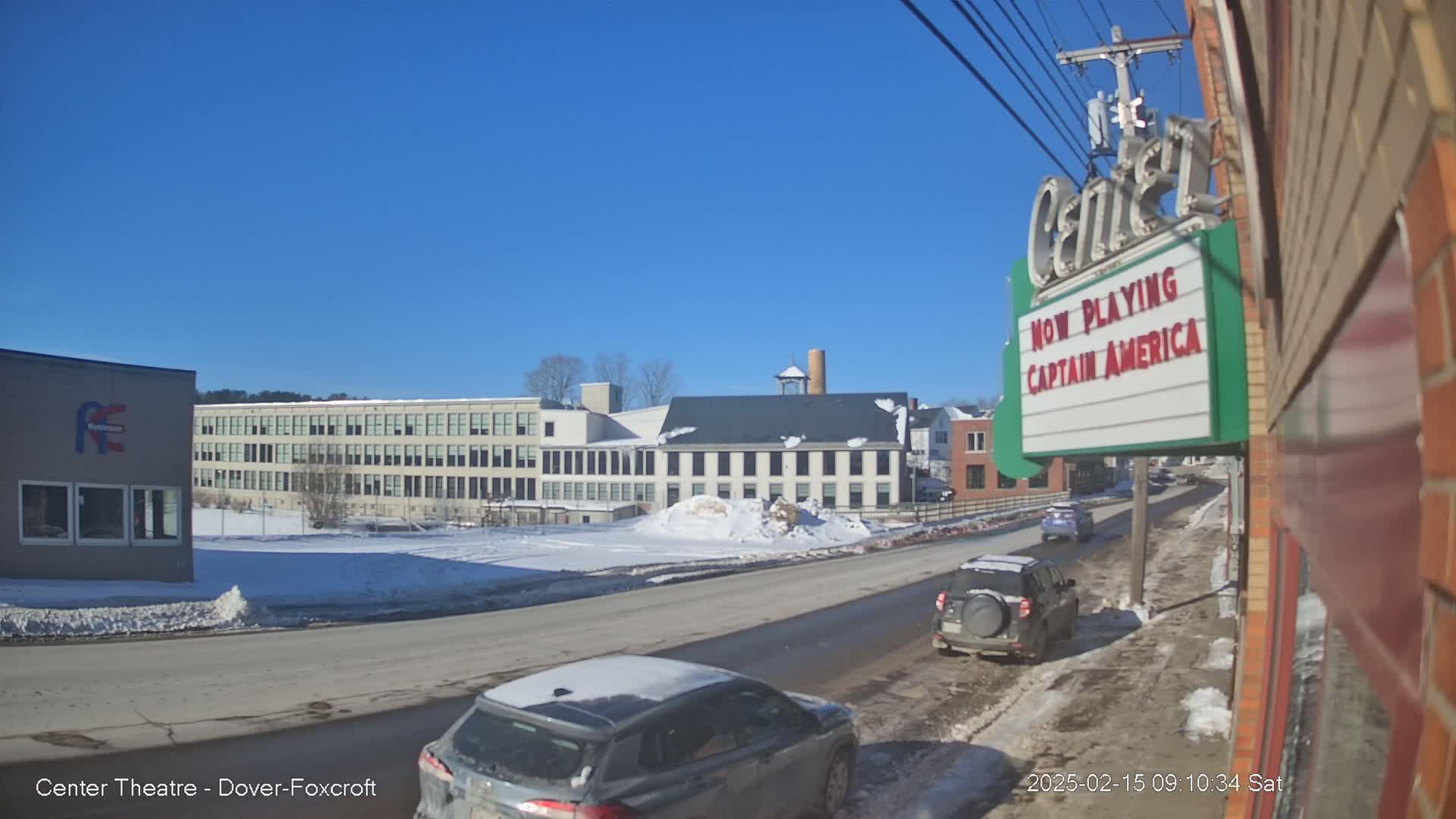 A sunny winter day shows a street scene with several cars, a snow-covered empty lot, and buildings including a large mill and a movie theater marquee advertising Captain America.