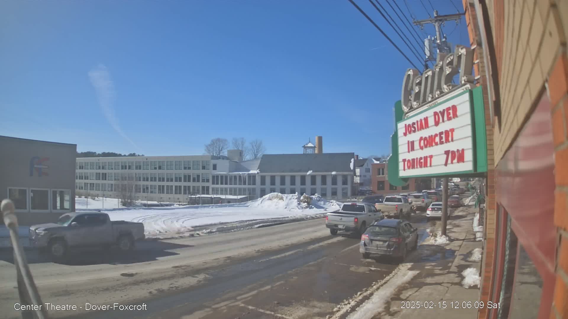 A partly sunny winter day shows a street scene with cars parked and driving past a large building, and a smaller building with a marquee advertising a concert.