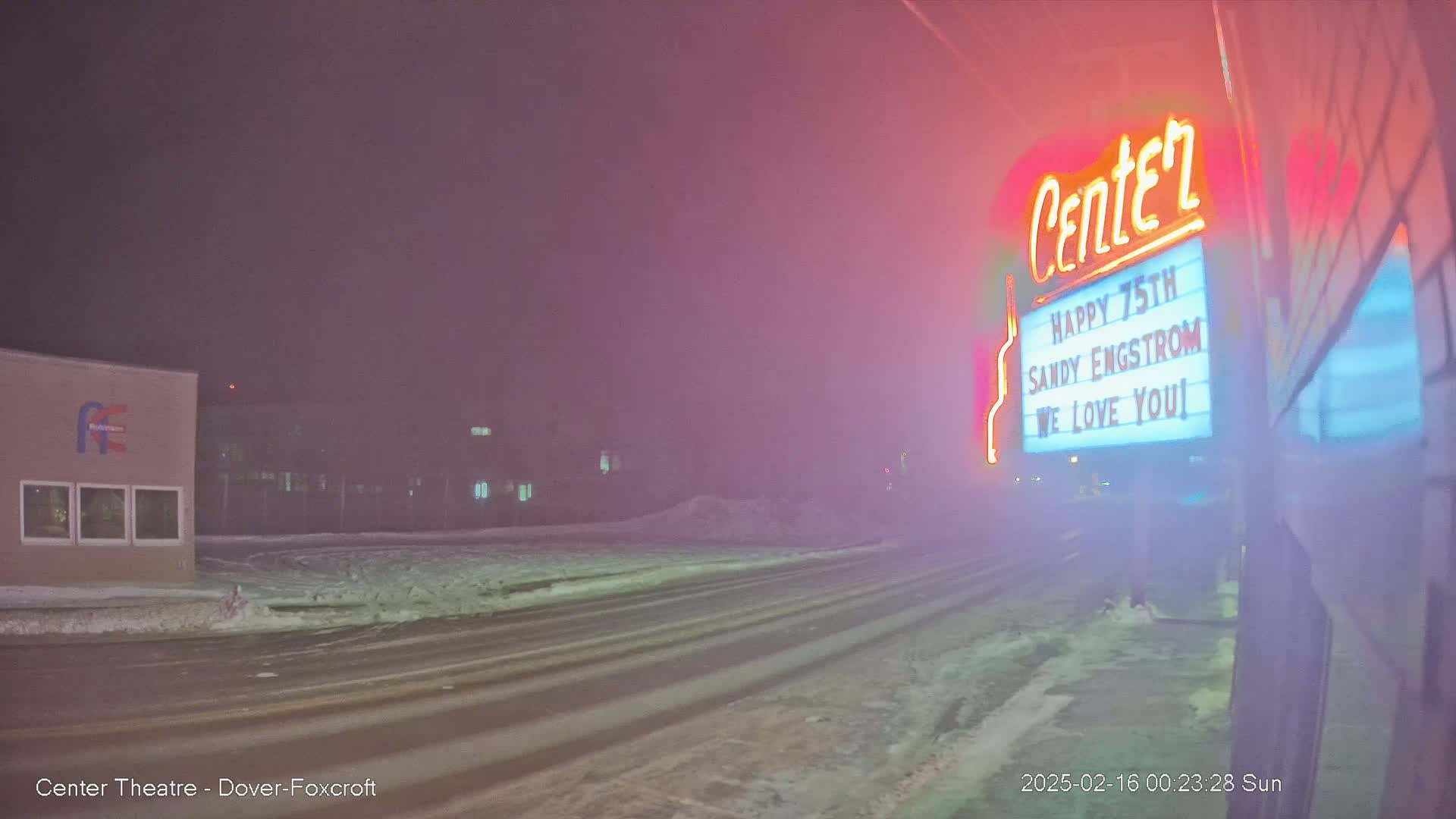 A snow-covered street at night is dimly lit by a neon sign on a building in foggy conditions.