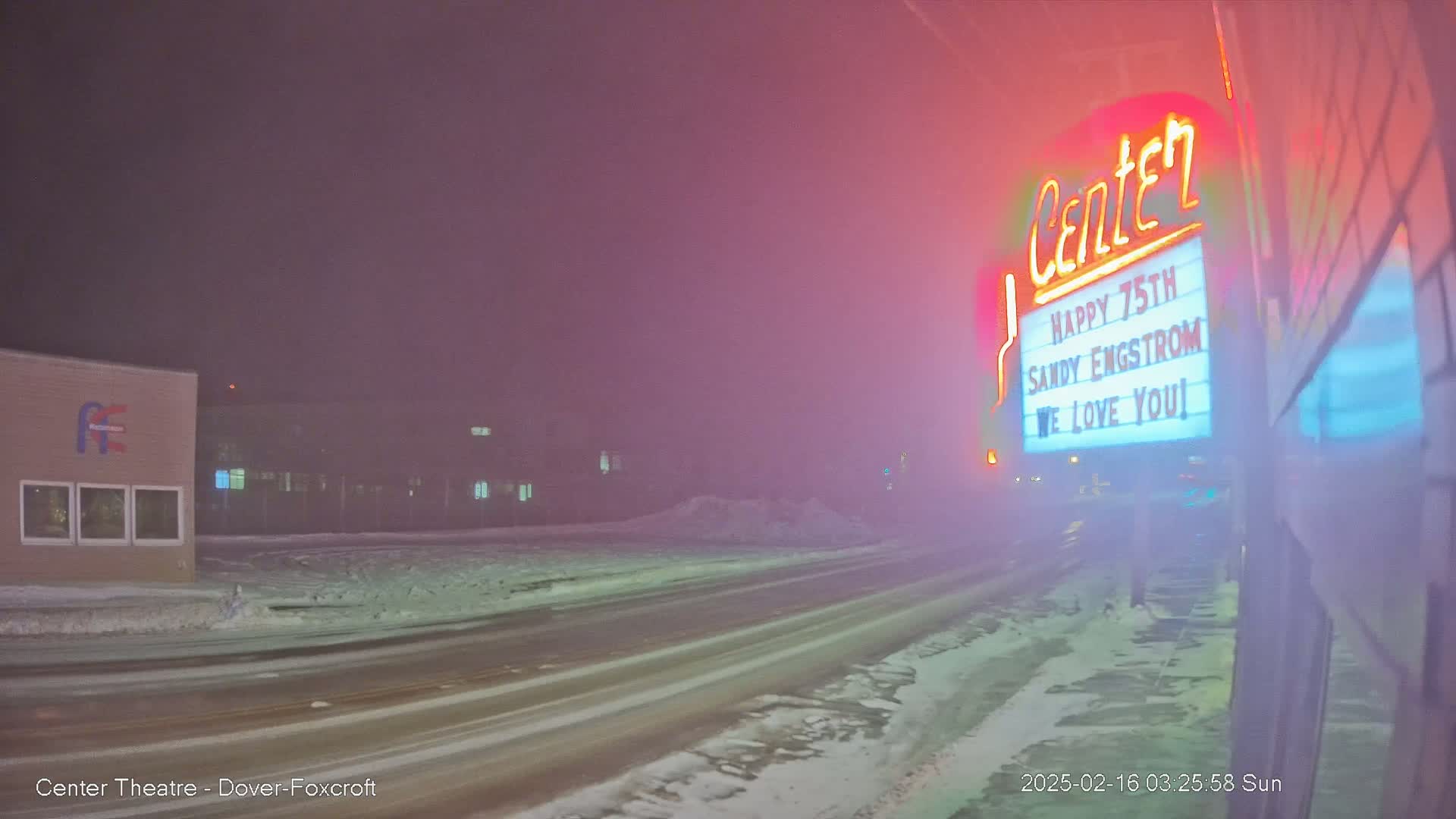 A snow-covered street at night is dimly lit by a neon sign on a building, with a hazy, foggy atmosphere.
