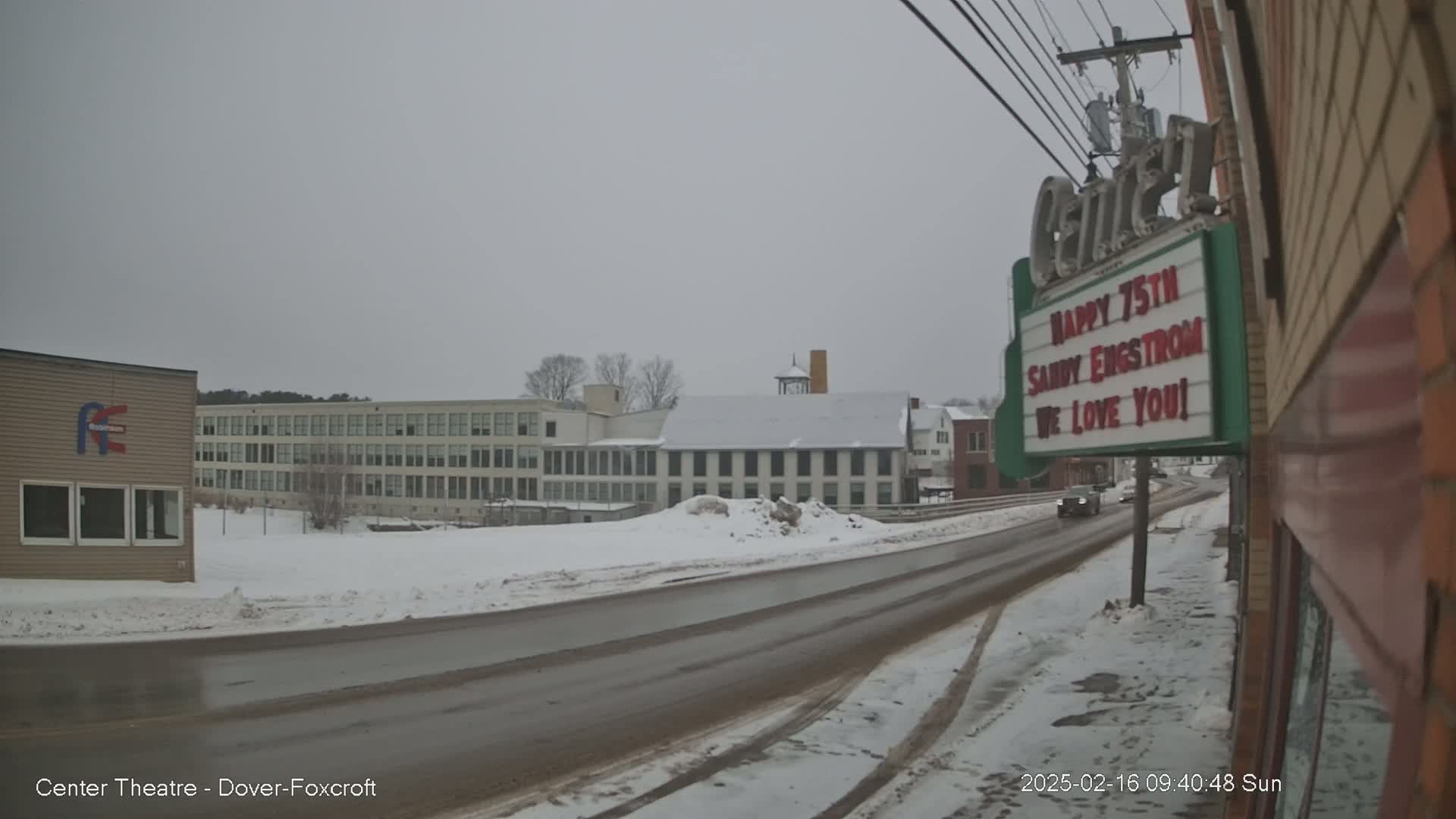 A snow-covered road leads past several buildings, including a large multi-story building and a smaller, snow-covered building, under an overcast sky.