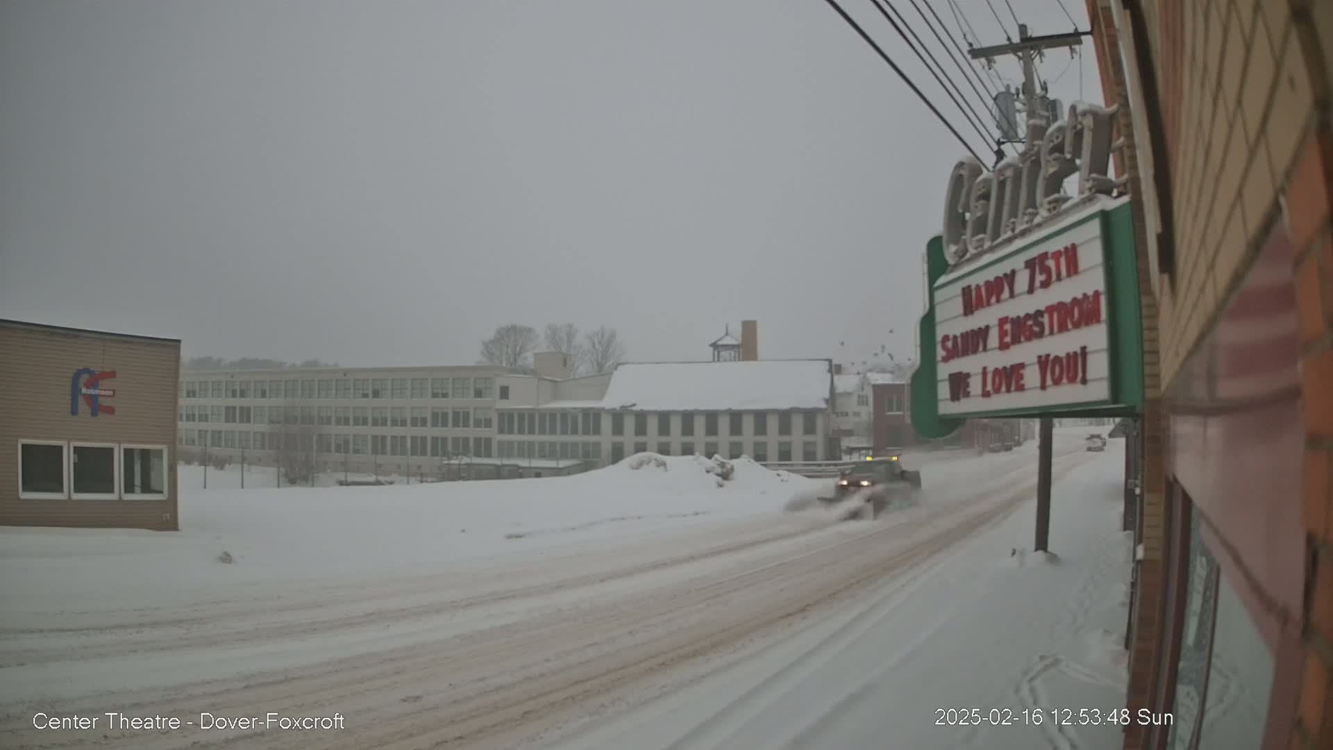 A snow-covered street scene shows a snowplow driving on a snow-covered road past buildings during a heavy snowfall.