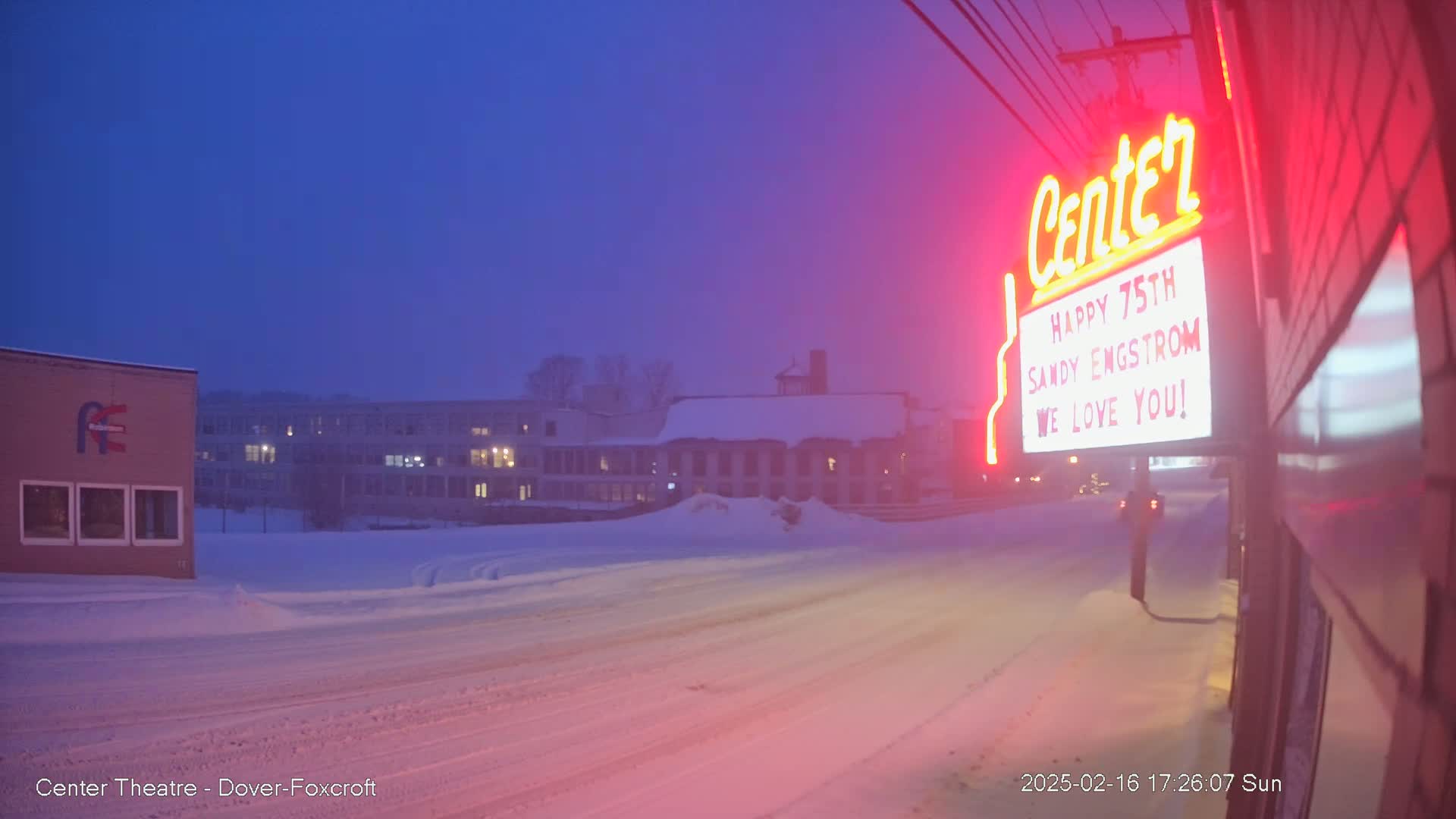 A snow-covered street scene at twilight shows a building with a neon sign celebrating a 75th birthday, with other buildings and snowdrifts visible in the background.