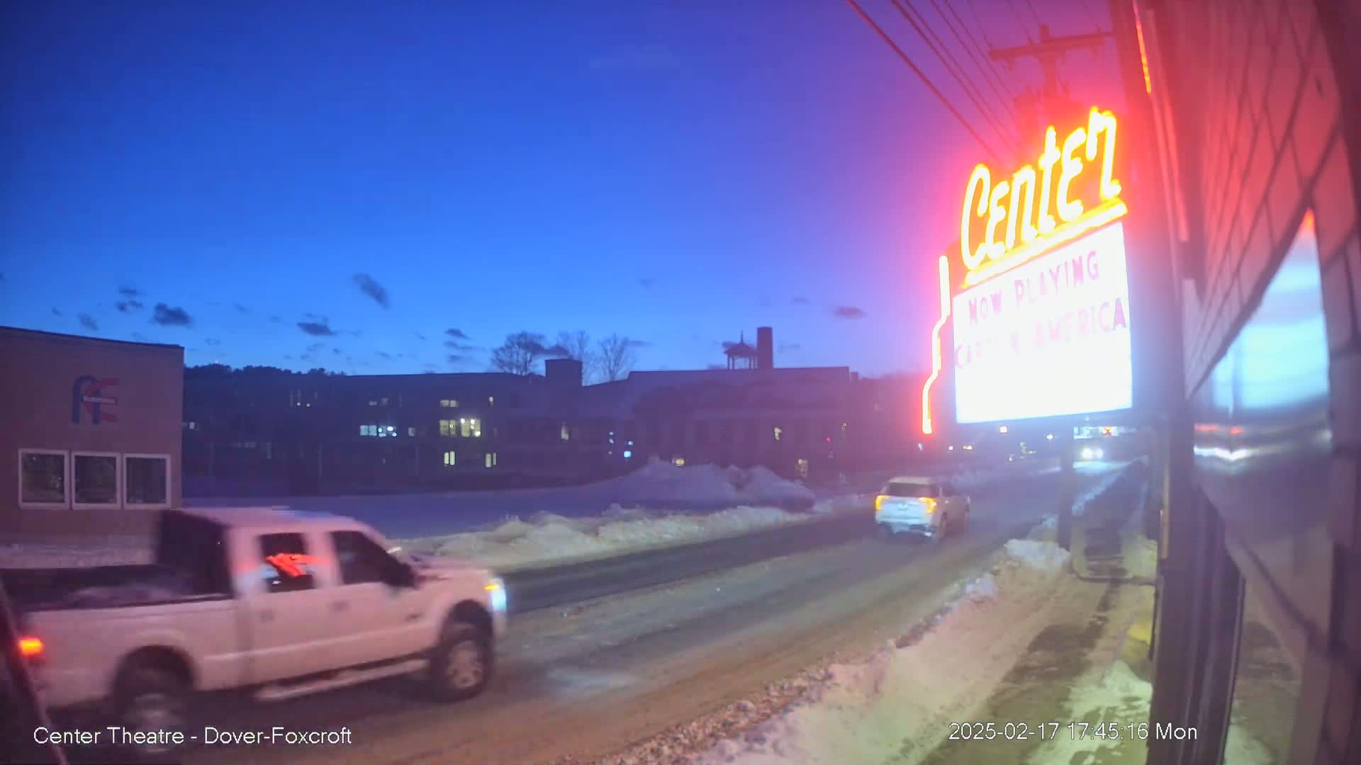 A white pickup truck drives past a movie theater marquee under a twilight sky, with snow visible on the roadside.