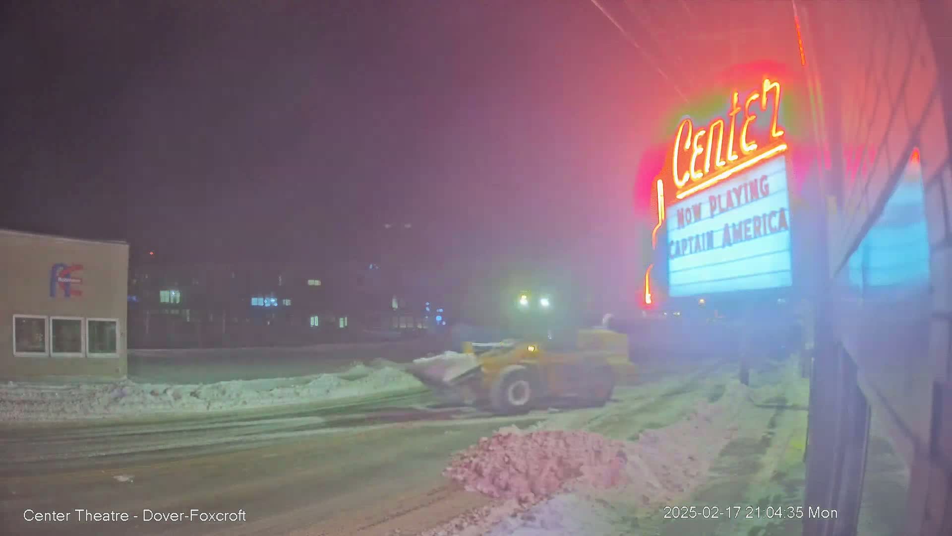 A snow plow clears a snow-covered street at night in front of a movie theater showing "Captain America."