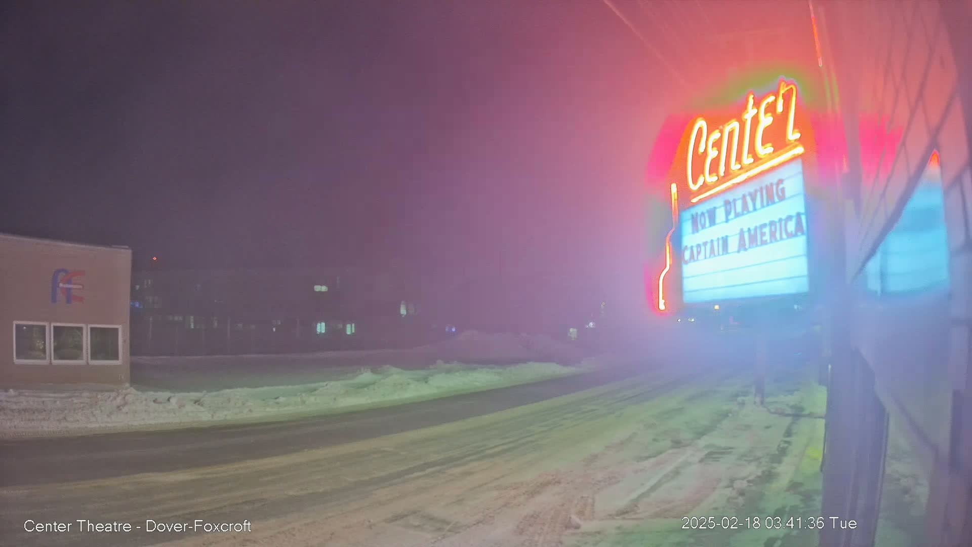 A snow-covered street at night is seen with a movie theater marquee displaying "Captain America" in the fog.