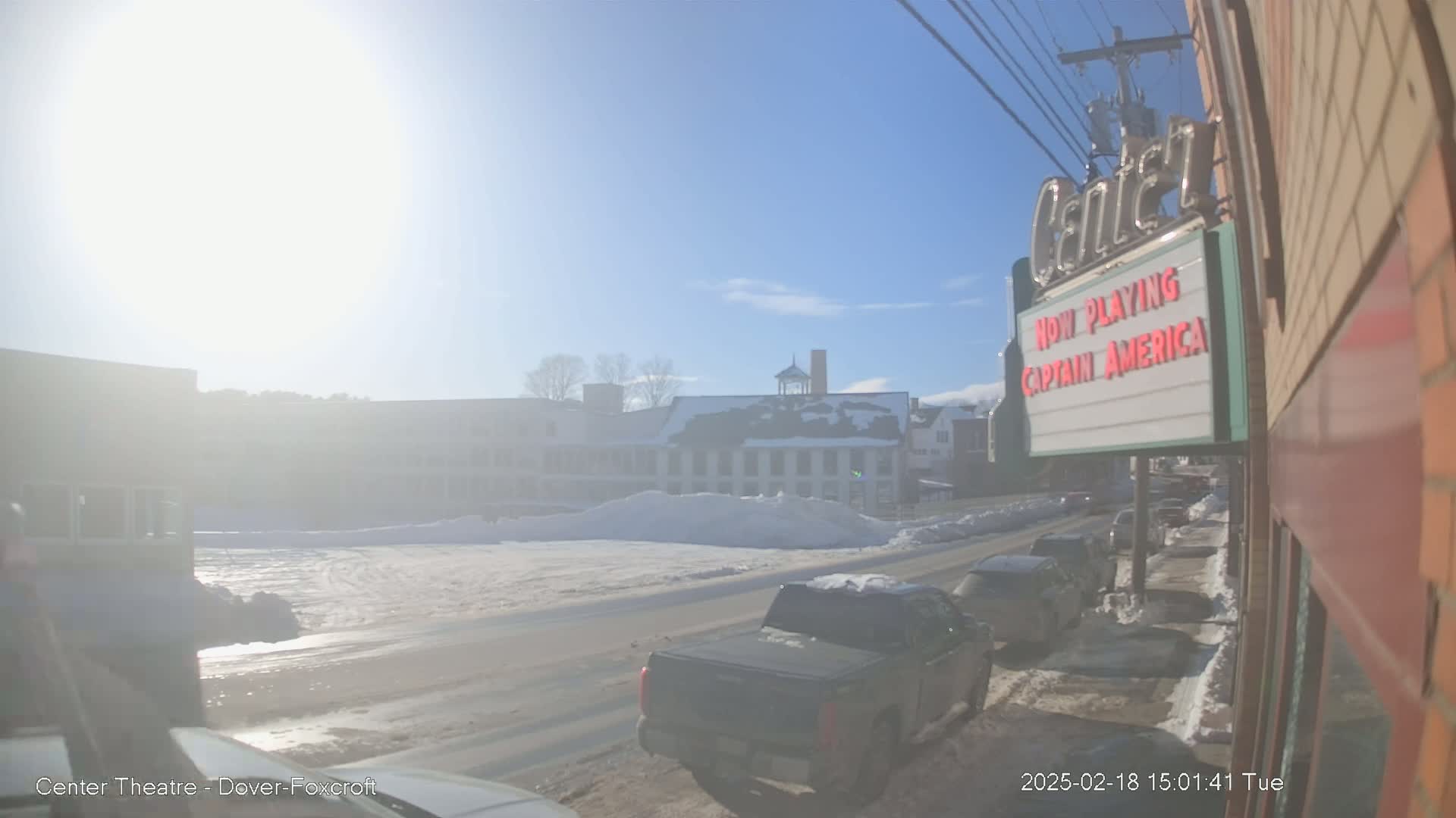 A sunny winter day reveals a snow-covered street with parked cars in front of a multi-story building, and a movie theater marquee advertising "Captain America".