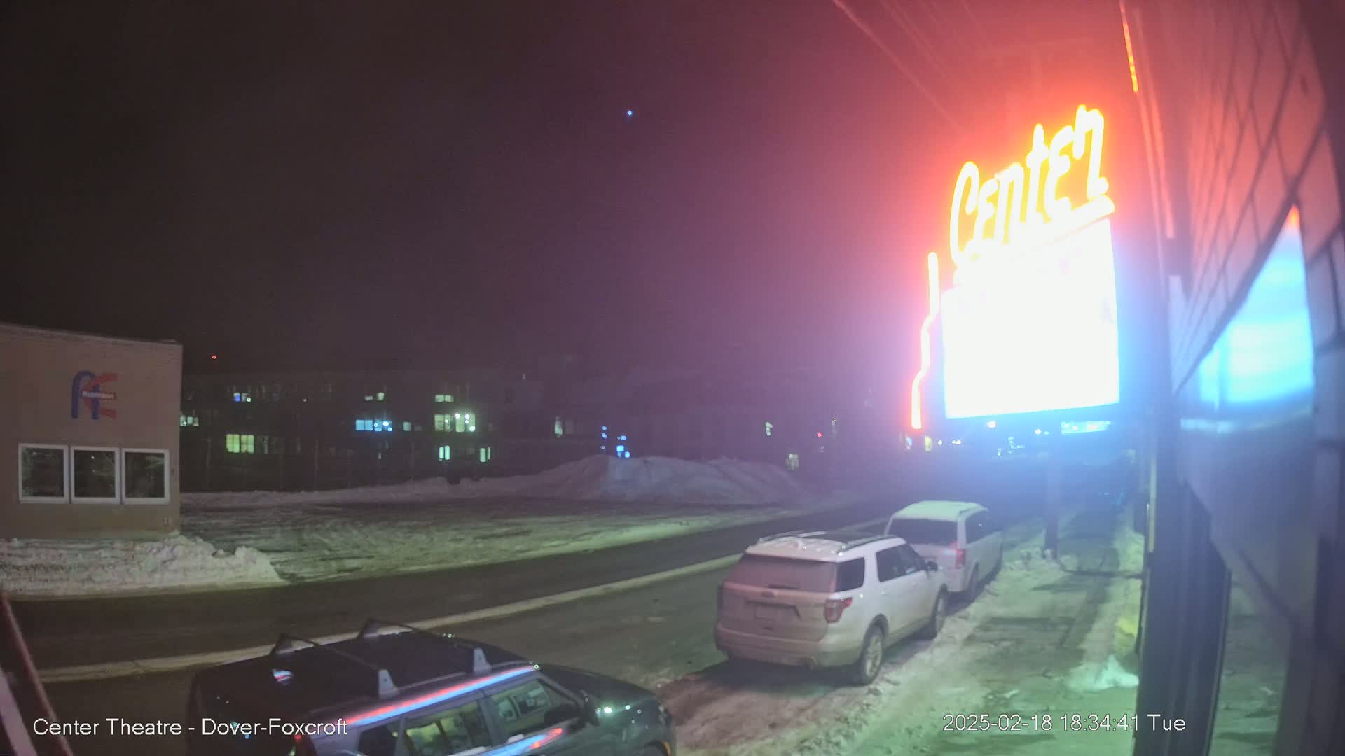 A nighttime street scene with snow on the ground shows two parked SUVs near a building with a neon sign.