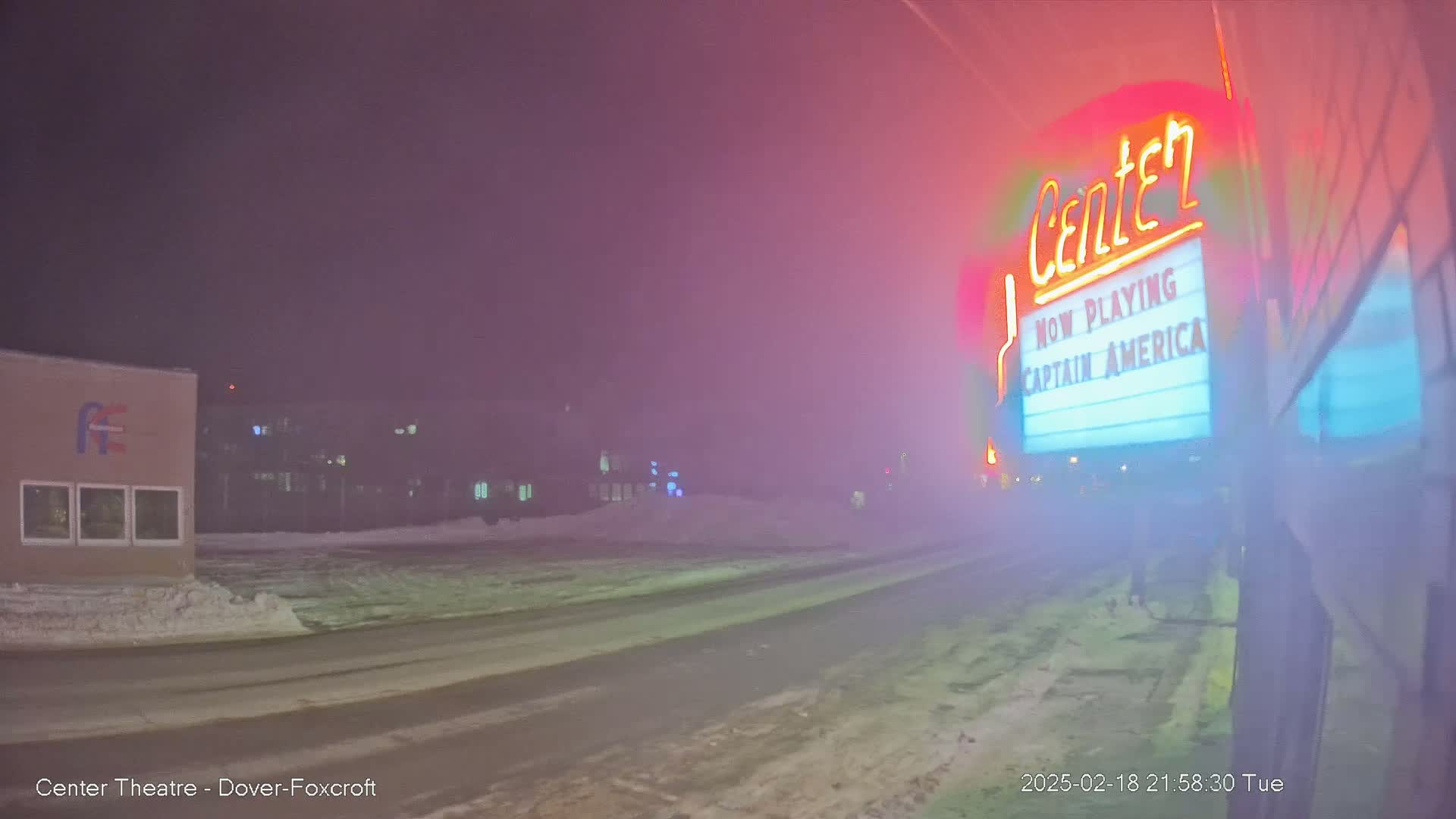 A snow-covered road at night leads to a movie theater marquee advertising "Captain America" under a hazy, pink-tinged sky.