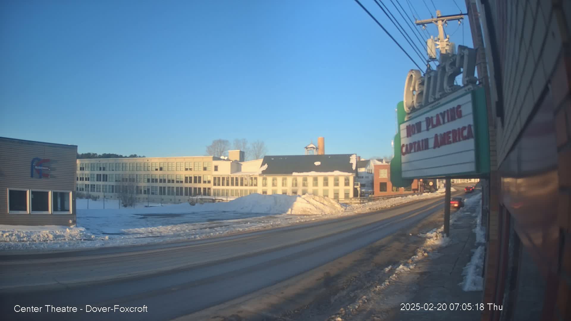 A sunny winter day reveals a snow-covered street scene with buildings, including a movie theater marquee advertising "Captain America," and several cars.