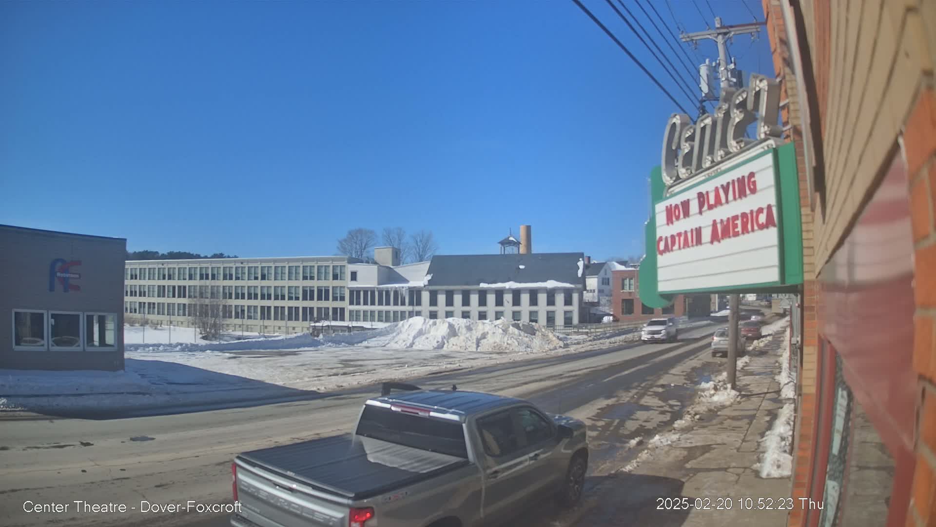 A street scene on a sunny winter day shows a pickup truck in the foreground, several cars driving down a road that passes in front of buildings including a large factory and a movie theater showing Captain America.