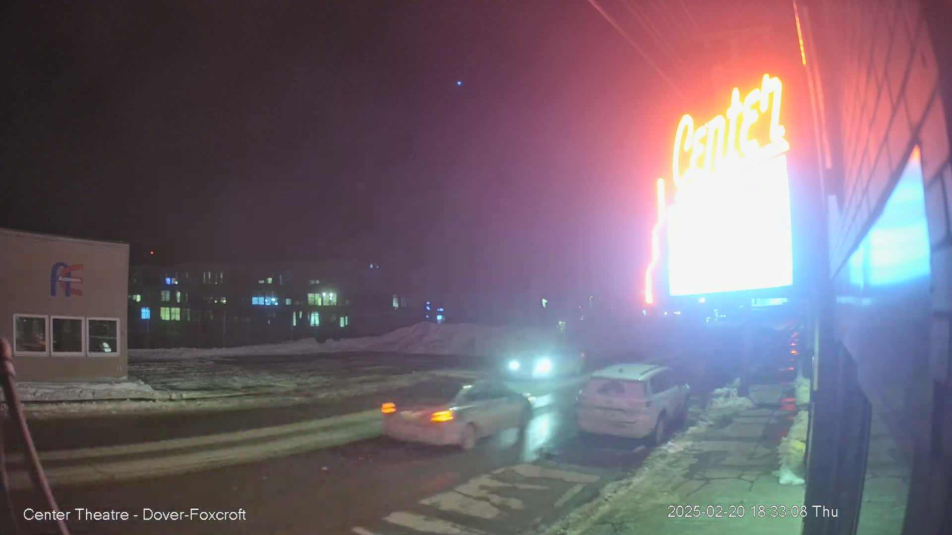 Two cars are parked near a building with a brightly lit sign at night in snowy conditions.