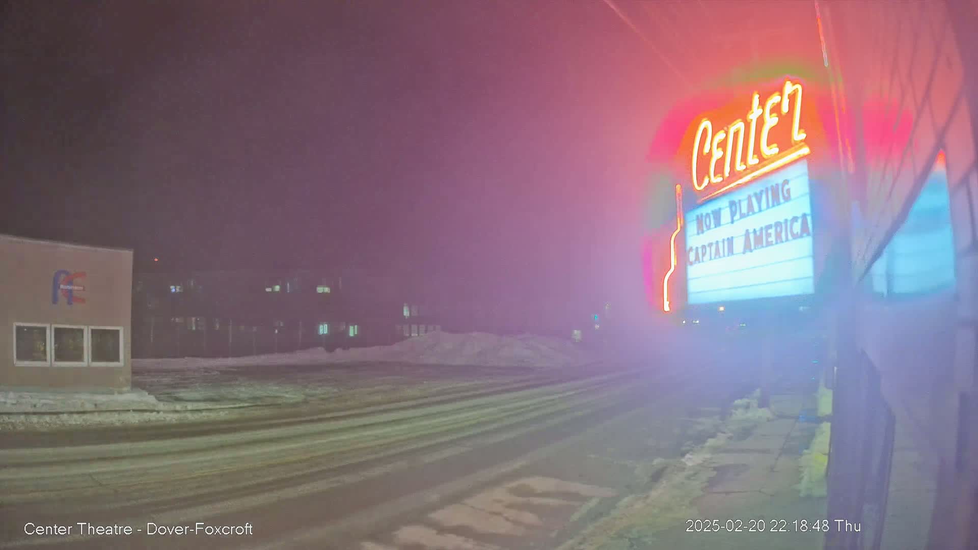 A snow-covered street at night is seen with a movie theater marquee displaying "Captain America" in the foreground, under hazy, possibly foggy conditions.
