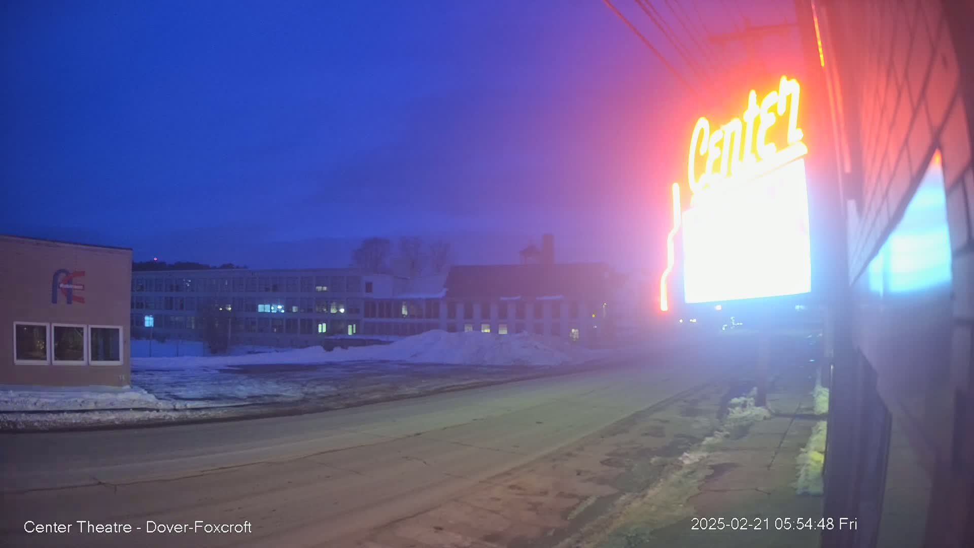 A brightly lit neon sign is partially visible on a building next to a snow-covered street at dusk, with other buildings in the background.