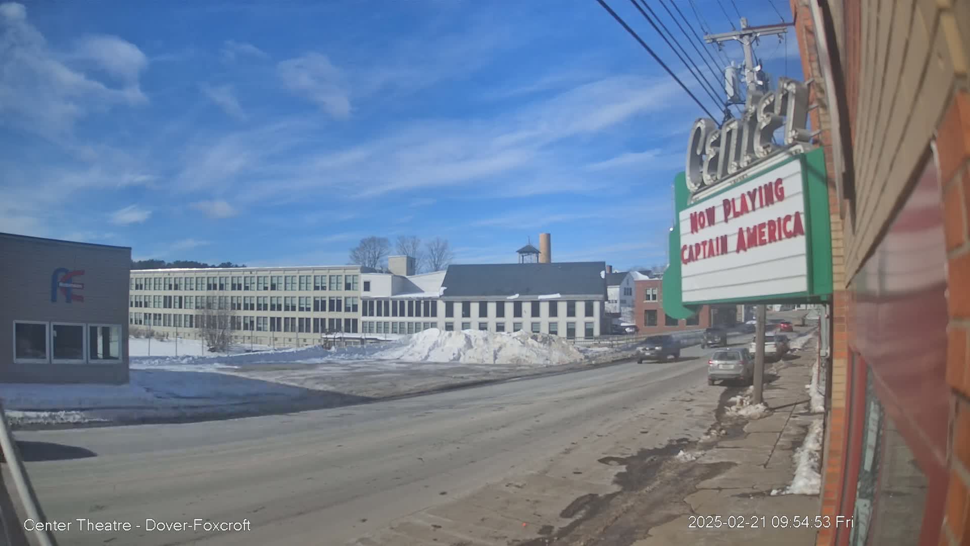 A street scene on a sunny day with a partly cloudy sky shows a snow-covered parking lot, several buildings, and cars driving on the road.